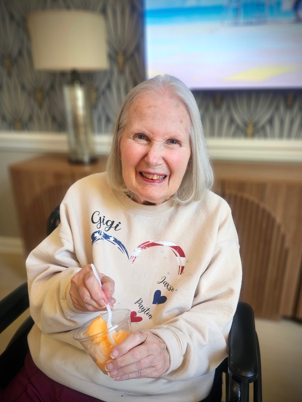 Older woman with gray hair smiling, holding a clear plastic bag with an orange object inside, sitting indoors.