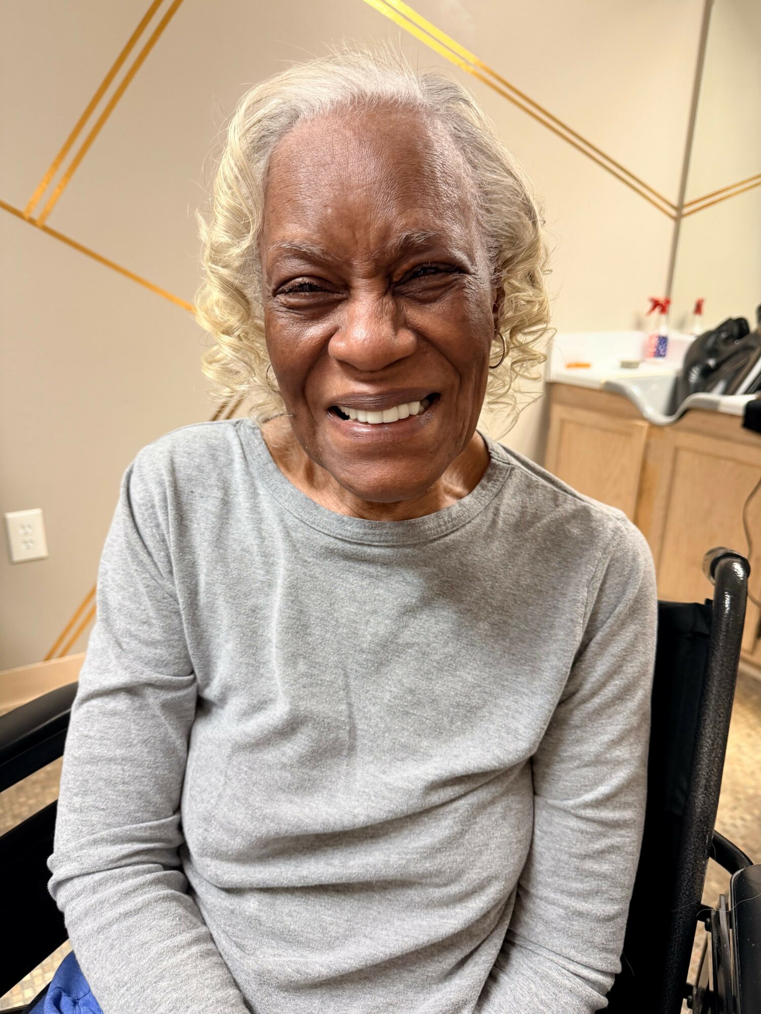 Elderly woman with curly gray hair smiling, wearing a gray long-sleeve shirt, sitting indoors.