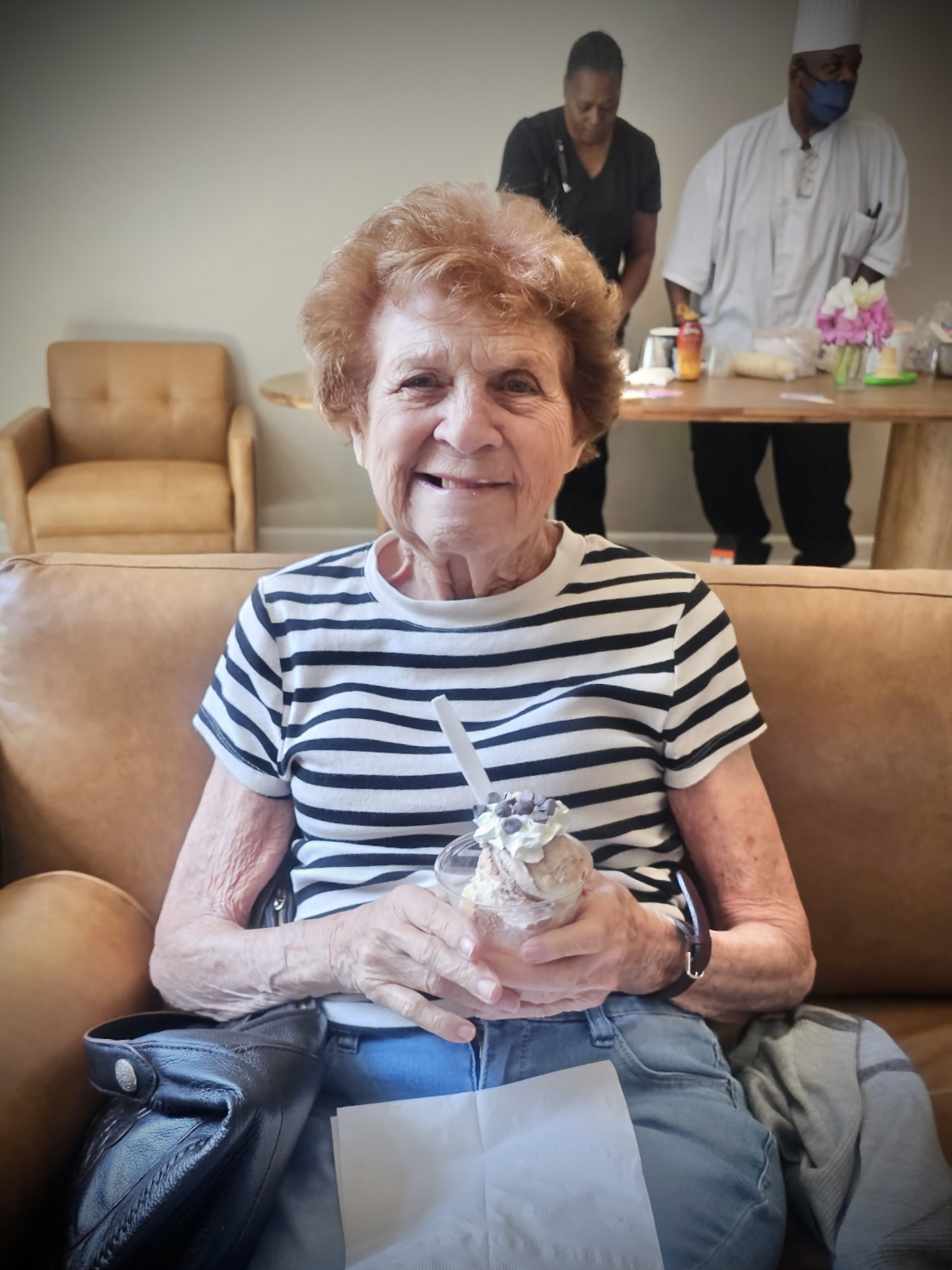 Elderly woman with short curly hair smiling, holding a wrapped gift, sitting on a sofa in a room with two people in the background.