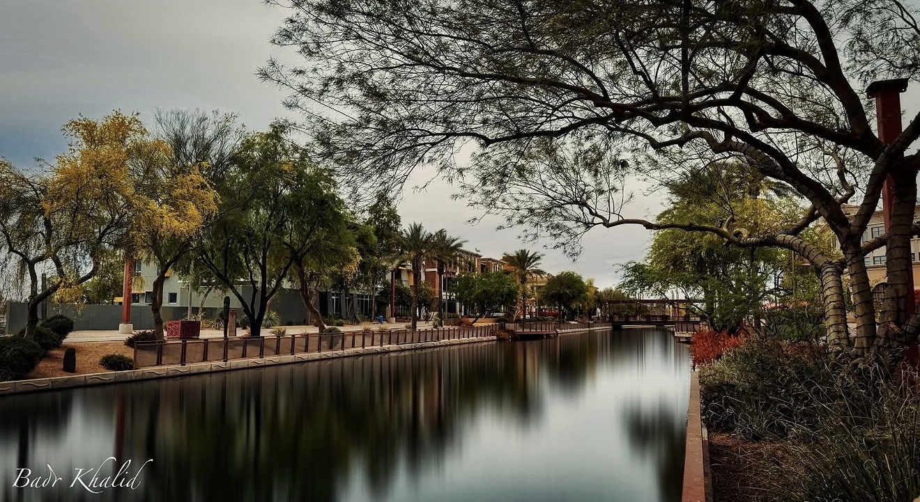 Calm waterway lined with trees and a walkway, with buildings and a bridge in the background.