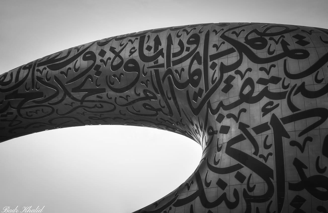 Close-up of a sculpture with Arabic calligraphy, featuring a curved surface and intricate black script on a gray background.