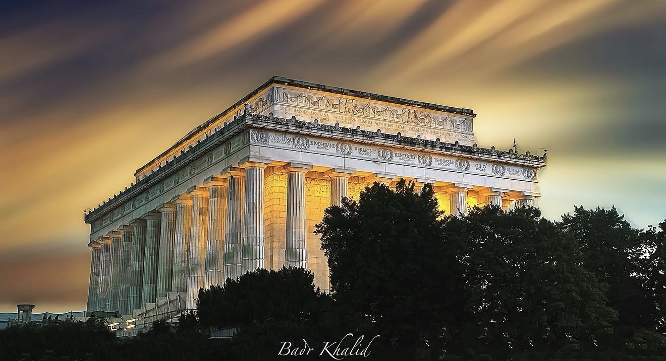 Ancient monument with columns illuminated at night, surrounded by trees and a colorful sky.