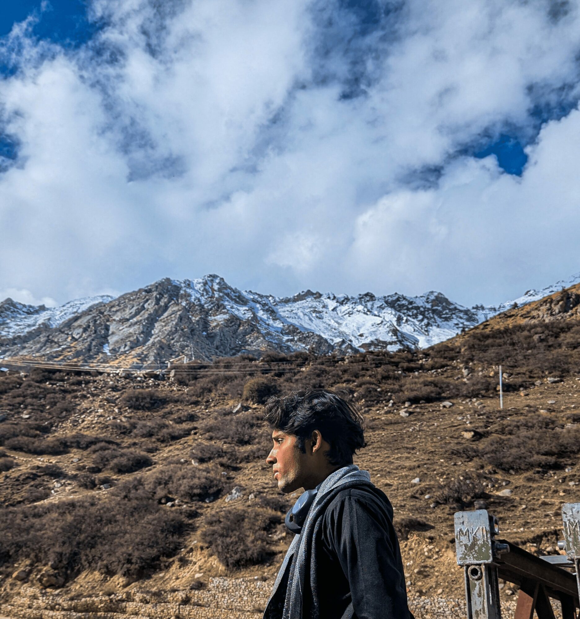 Young man with dark hair in profile, wearing a dark jacket, standing outdoors with mountains and sky in background.
