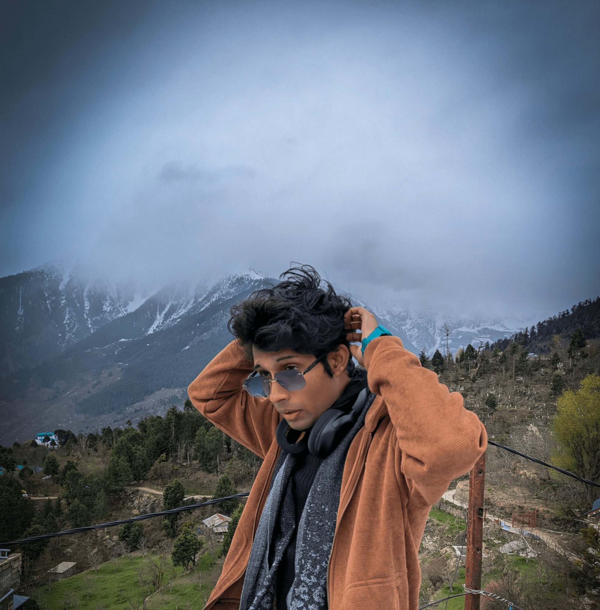 Young person with dark hair wearing sunglasses, brown jacket, and scarf outdoors with mountains and cloudy sky.