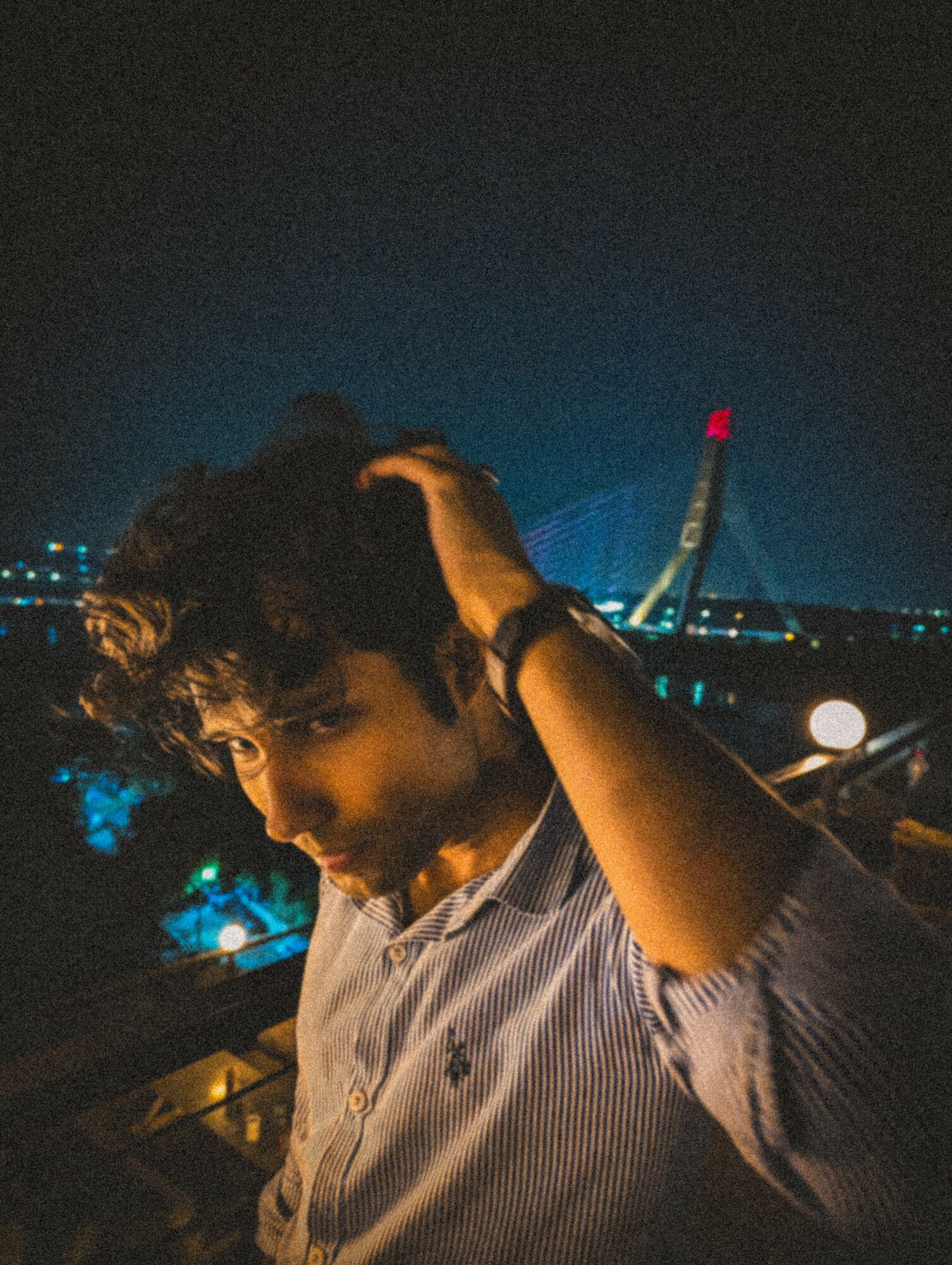 Person with curly hair adjusting hair outdoors at night, city lights and a bridge in the background.