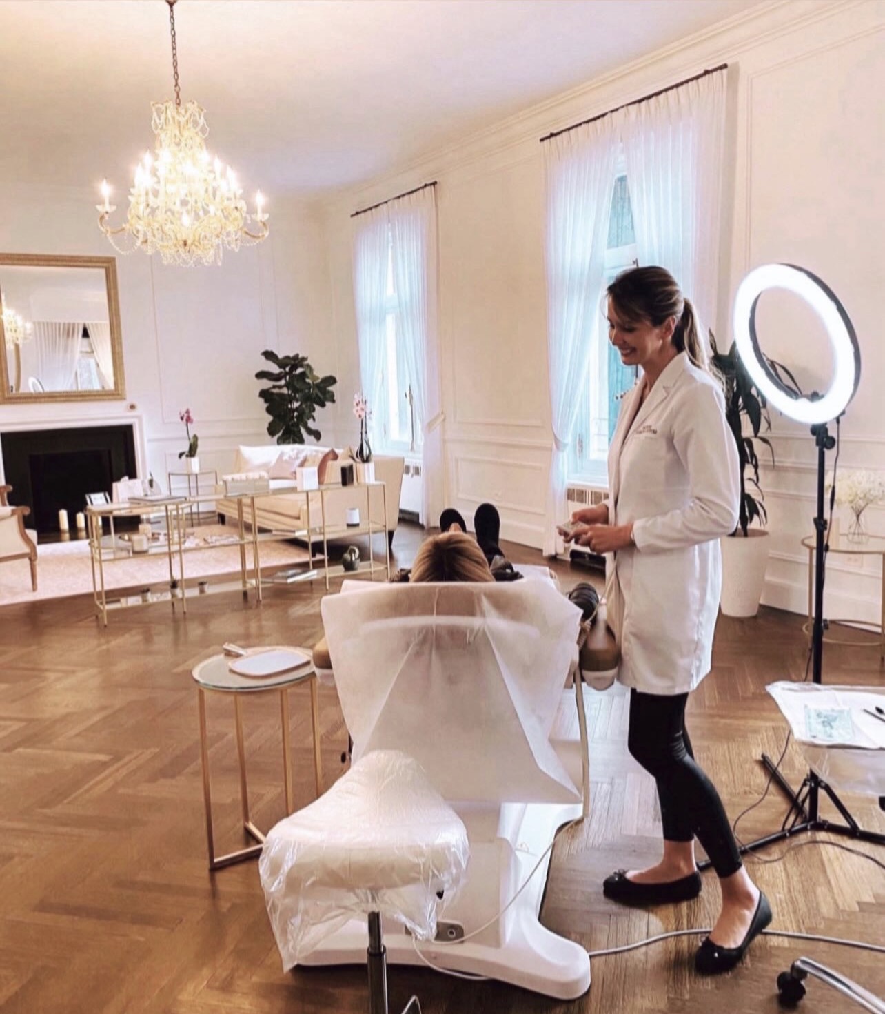 A woman in a white coat stands in a bright, elegant room with a ring light and a person seated at a table.