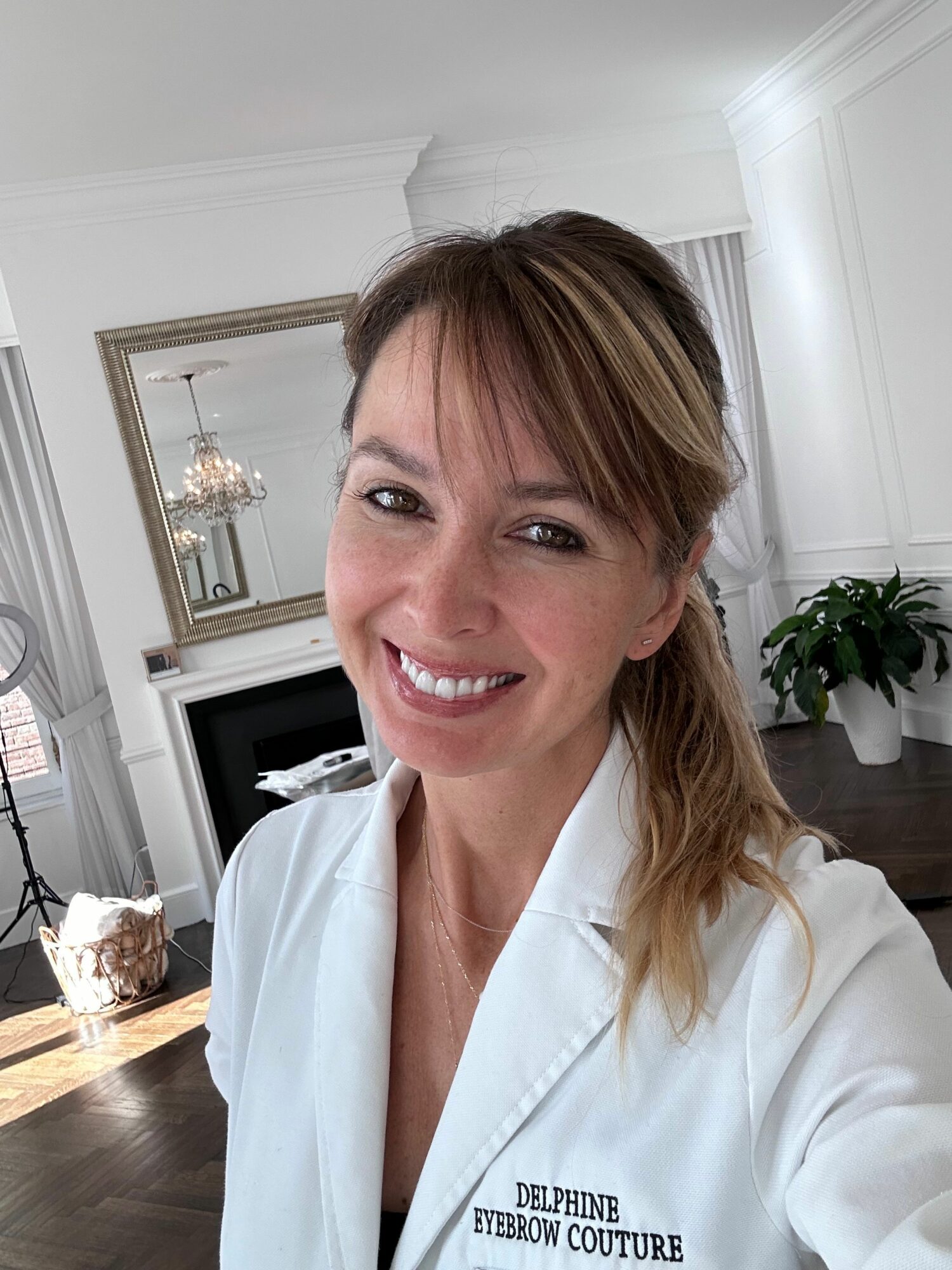 Woman with light brown hair smiling in a white coat in a room with a mirror and fireplace.