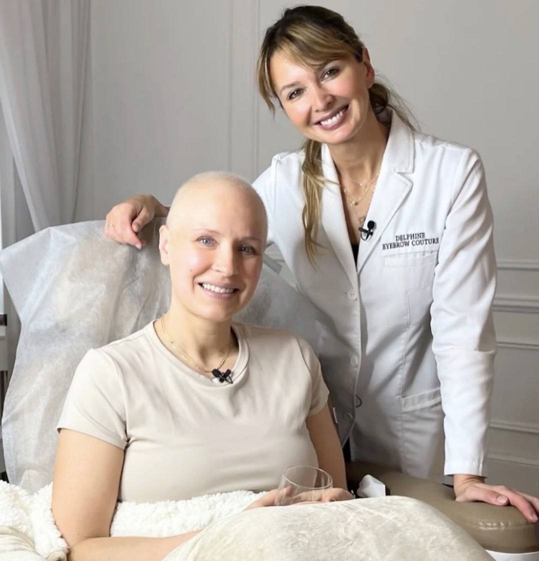 A woman with a shaved head sitting in a medical setting, smiling, with a healthcare professional standing beside her.