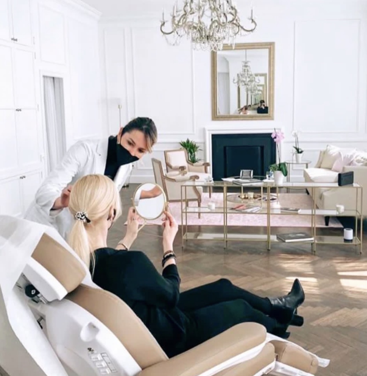 Woman sitting in a chair holding a mirror, woman standing nearby in a bright, elegant living room with chandelier and mirror.