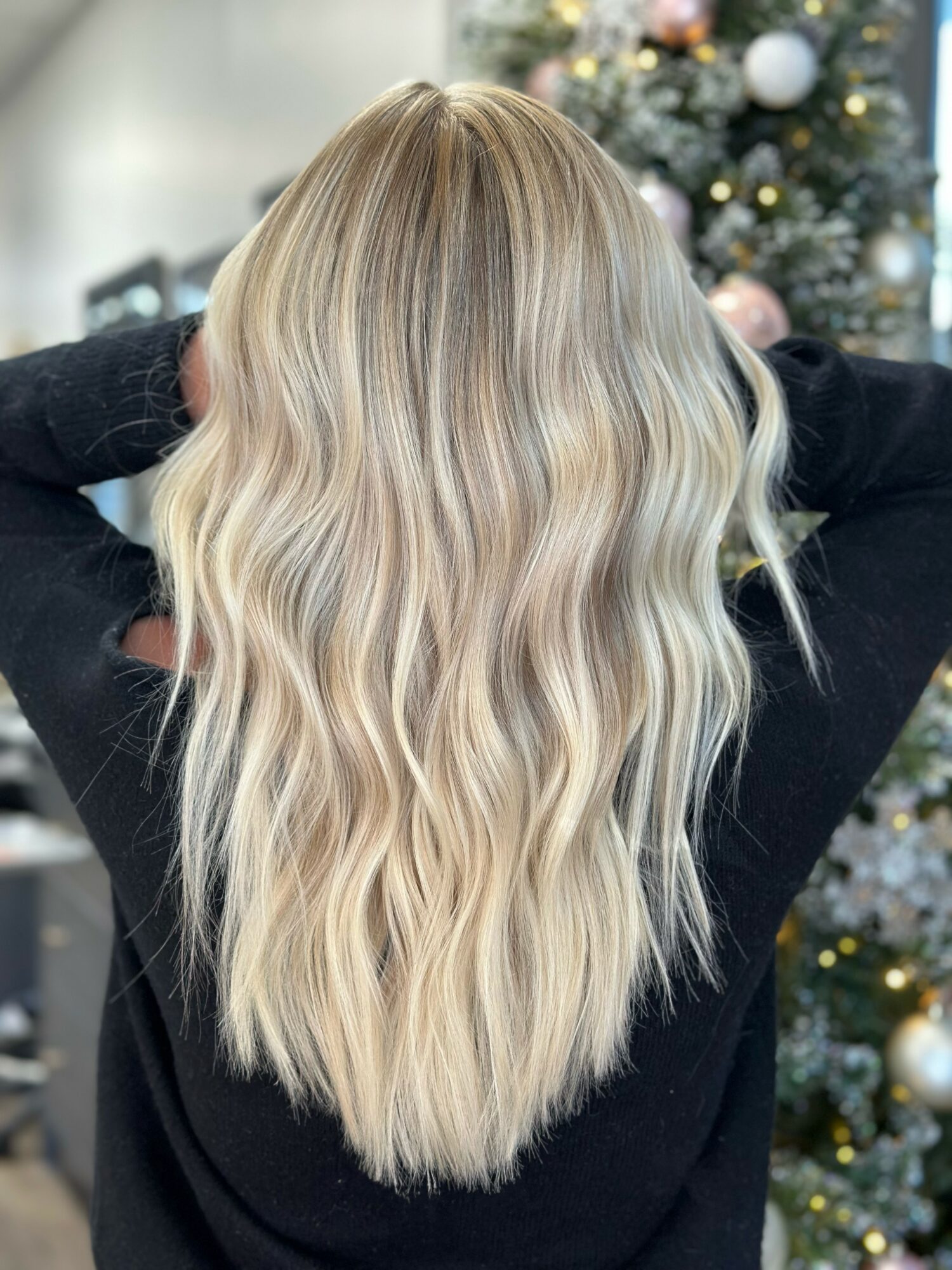 Back of a person with long, wavy blonde hair, standing in front of a decorated Christmas tree.