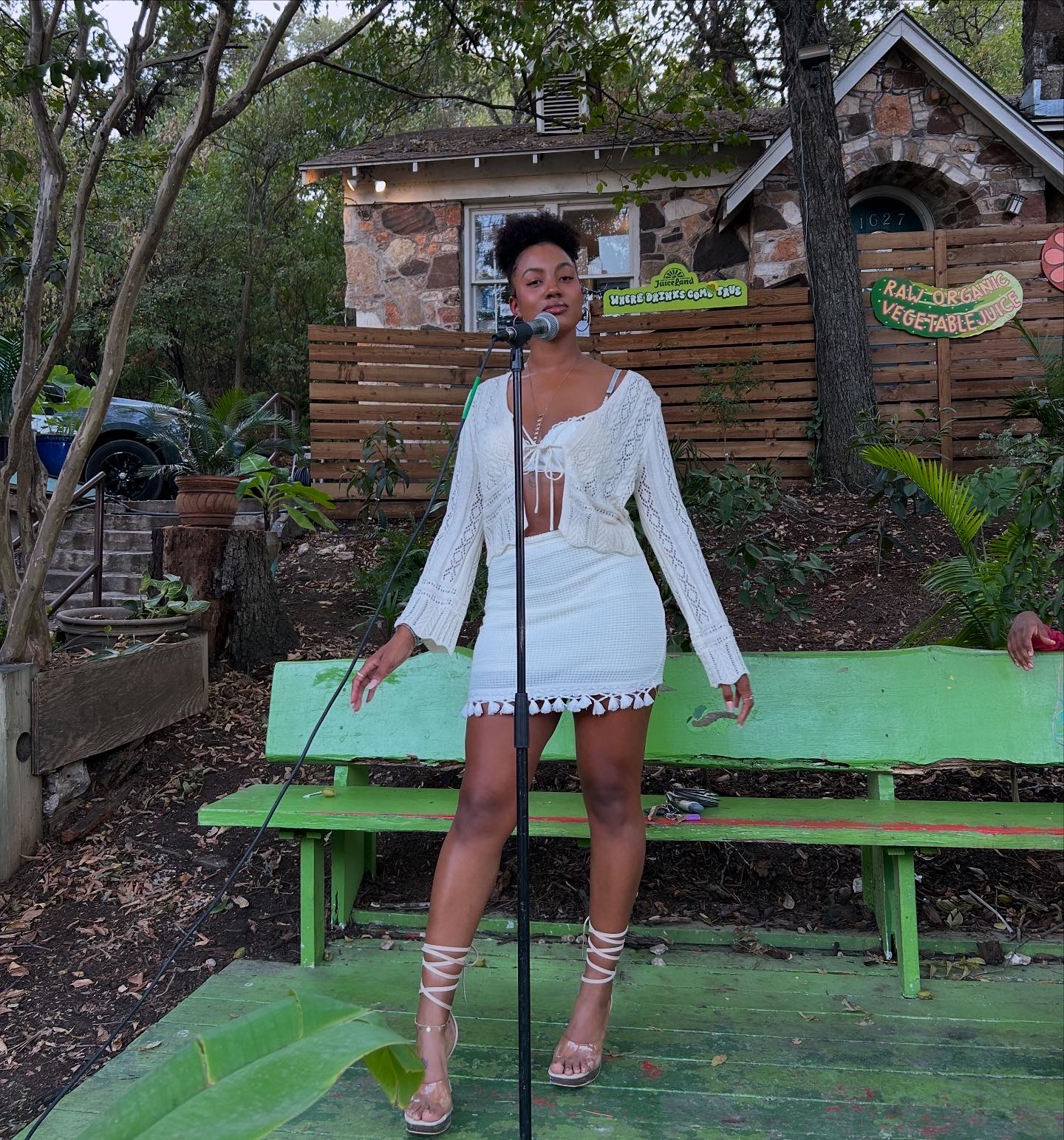 Woman standing outdoors in front of a green bench, wearing a white dress and heels, with trees and a building behind.