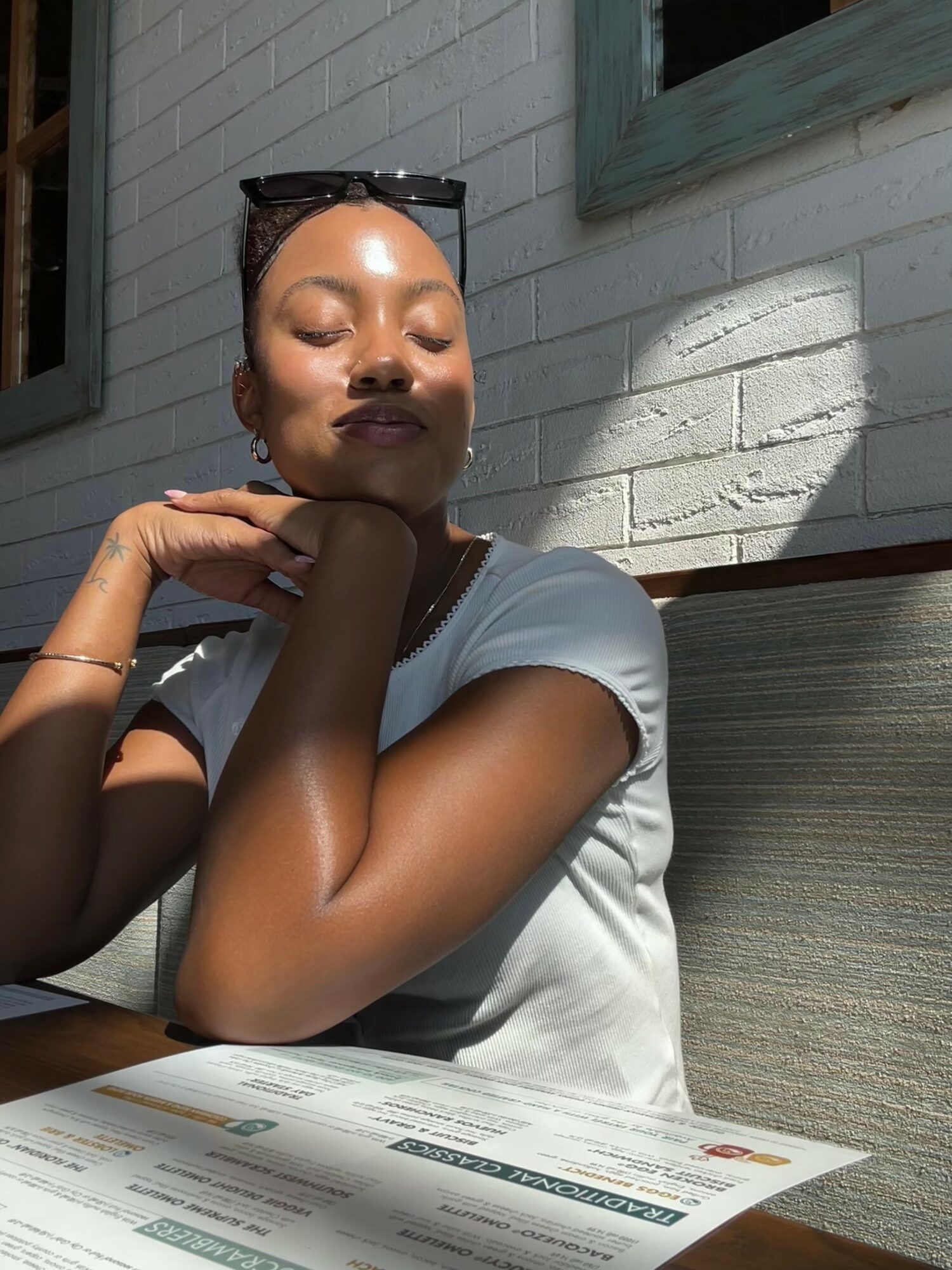 Woman sitting at a table outdoors with sunlight on her face, wearing sunglasses on her head and a white shirt.