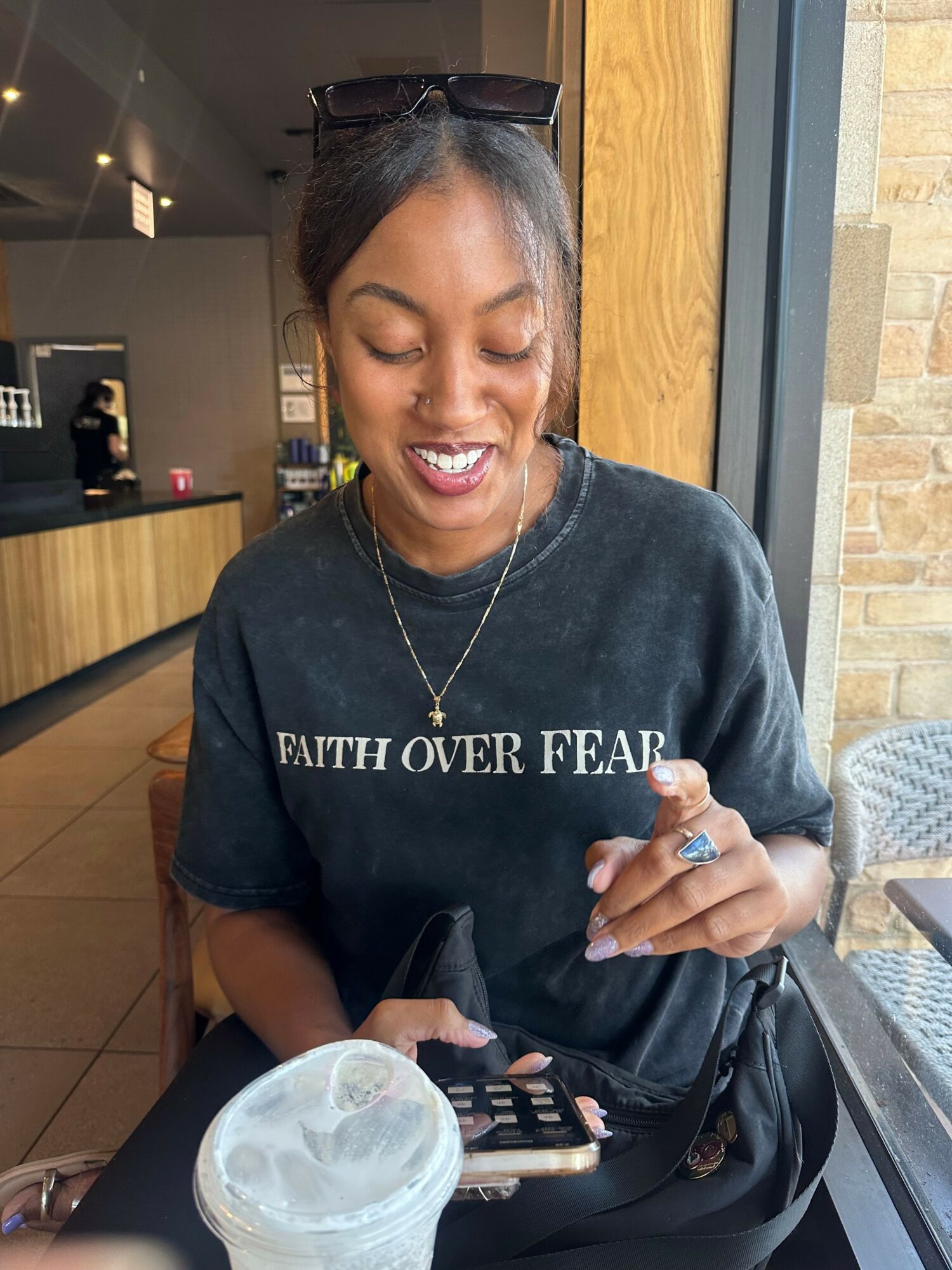 Young woman with sunglasses on her head, smiling, sitting at a table with a drink and phone, wearing a black T-shirt with text.