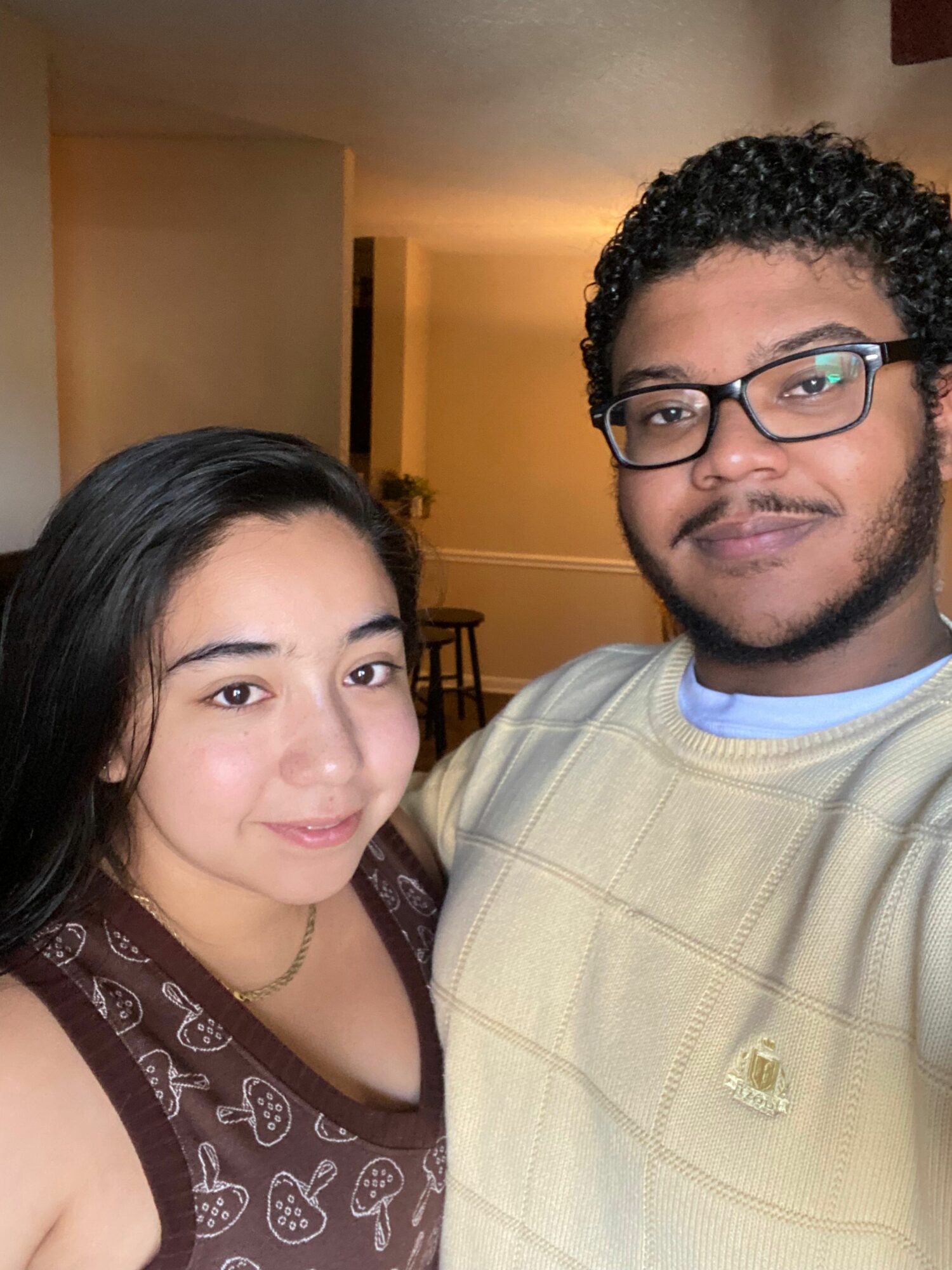 A man and woman pose indoors, smiling at the camera, with warm lighting and a blurred background.