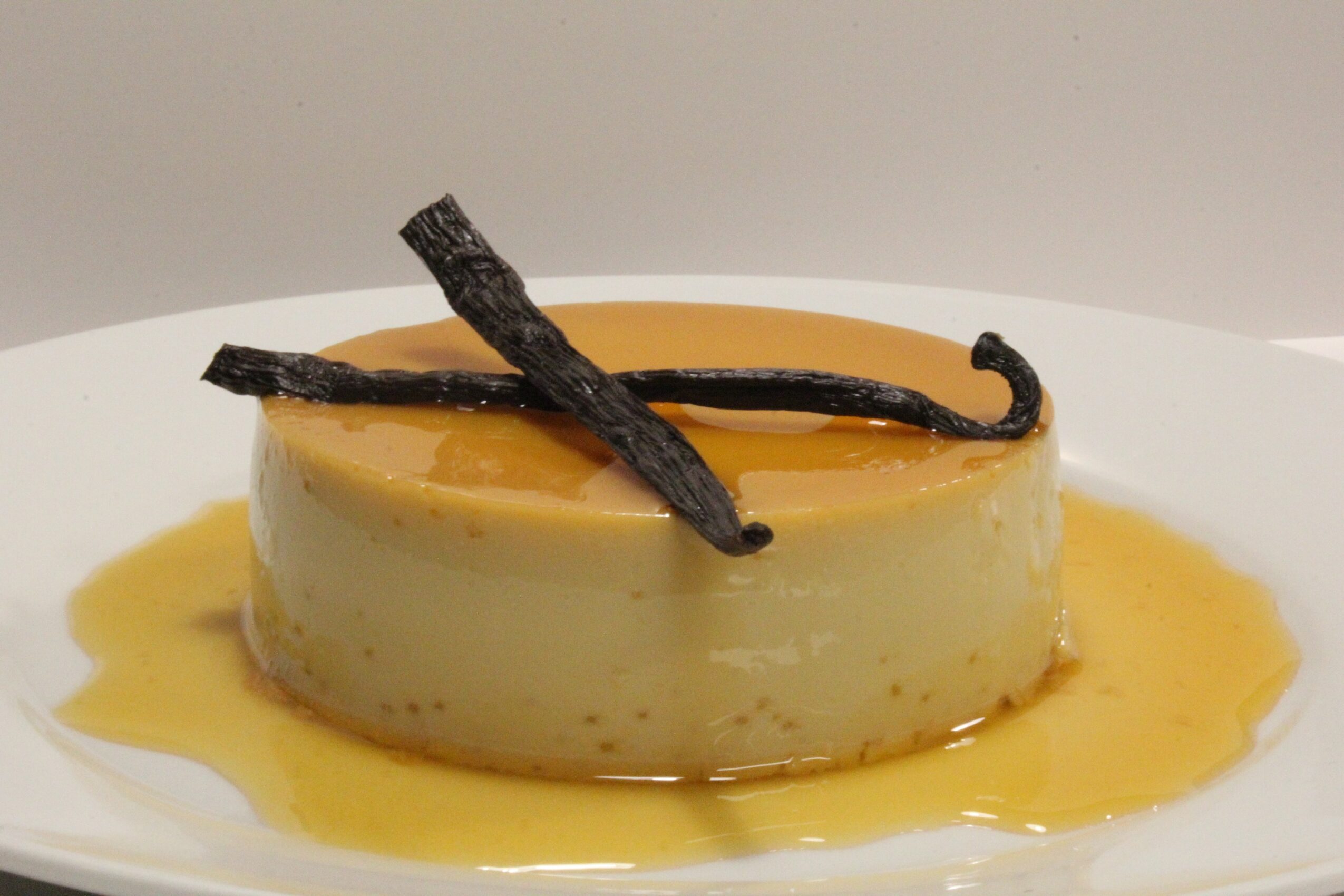 Round dessert with caramel sauce and two vanilla beans on top, served on a white plate.
