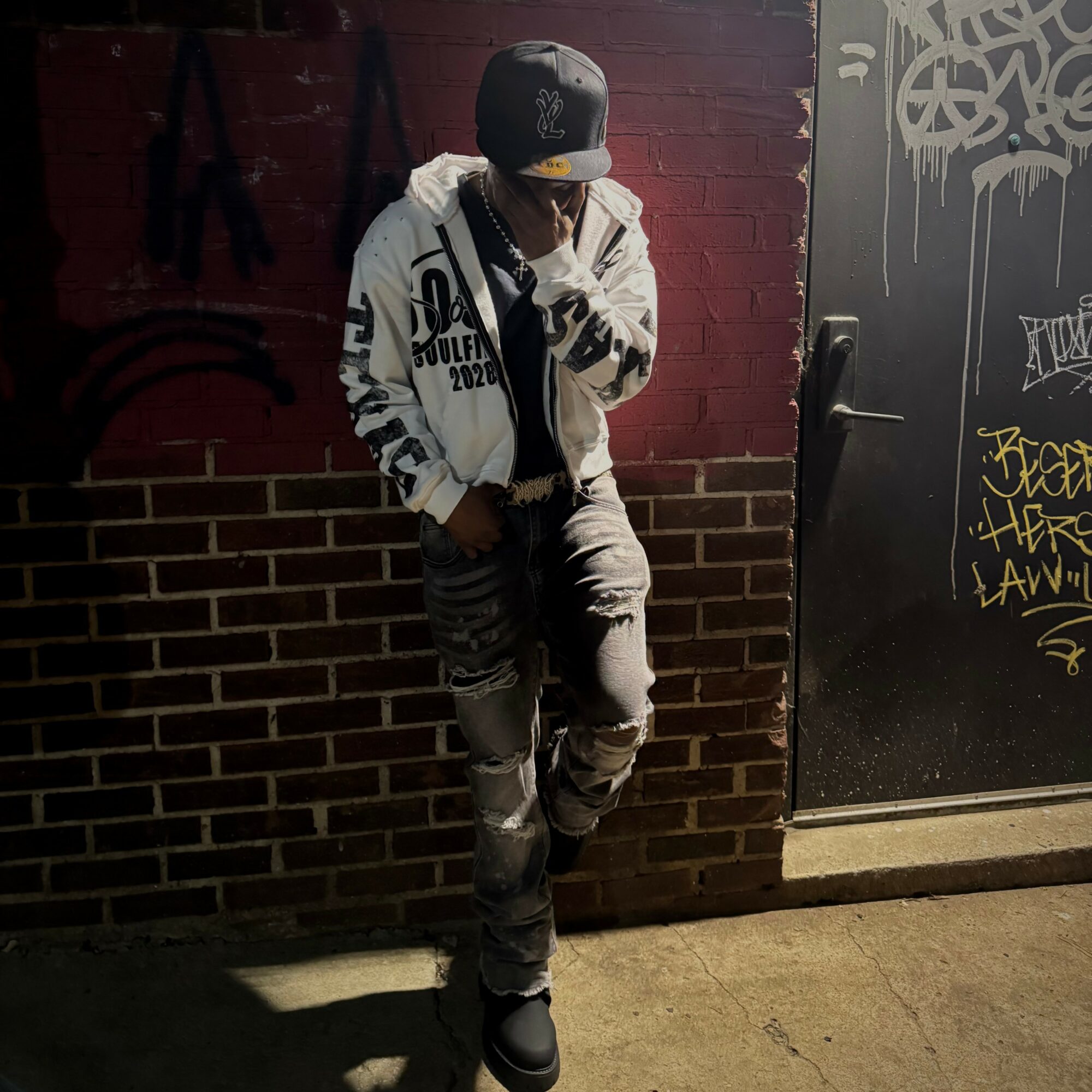 Person leaning against a brick wall, wearing a cap, hoodie, and ripped jeans, near a graffiti-covered door.