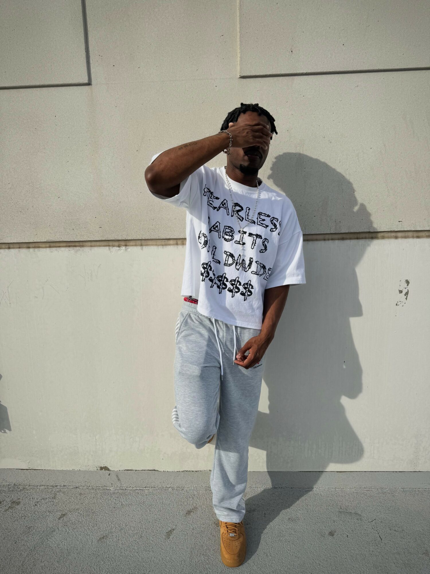 Young man standing against a plain wall, wearing a white graphic t-shirt, light jeans, and tan shoes, with one hand on his head.