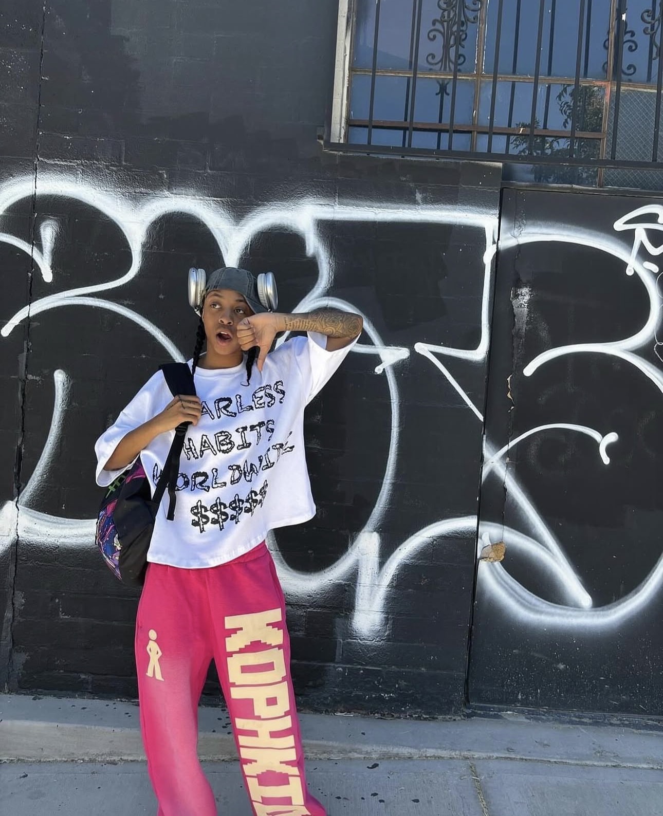 Person standing in front of graffiti-covered wall, wearing pink pants and a white t-shirt, holding a bag and making a gesture.