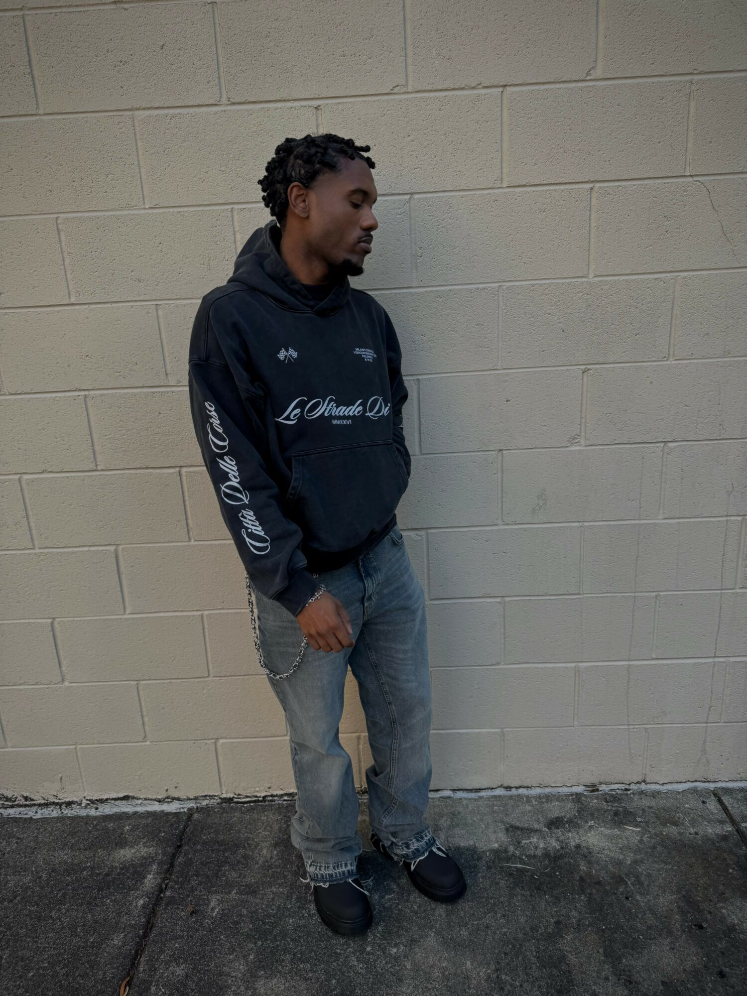 Young man with curly hair stands against a beige brick wall, wearing a black hoodie and jeans, looking to the side.