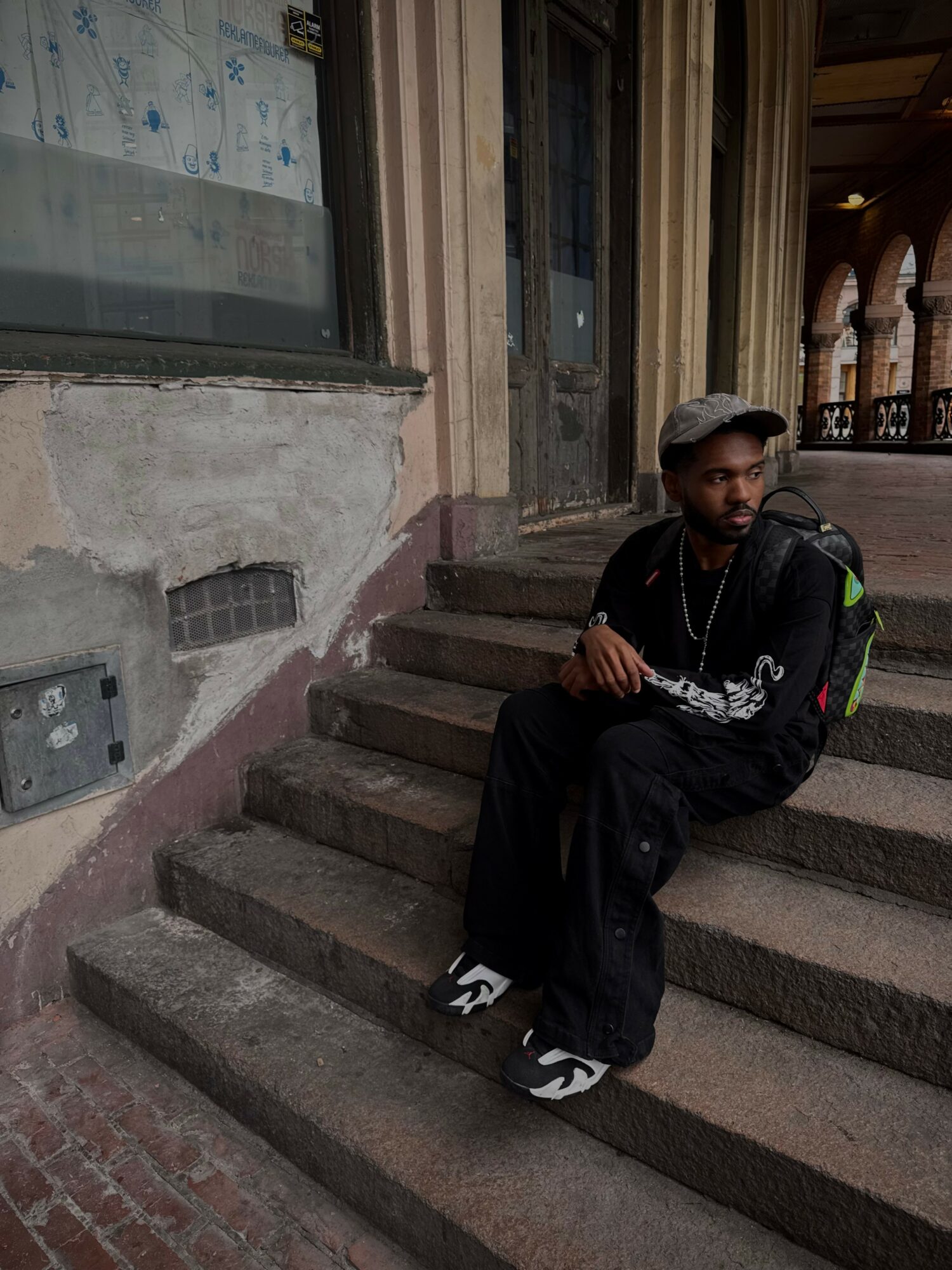 Man sitting on stairs outside building with large windows and wooden arches, wearing a cap, black jacket, and sneakers.
