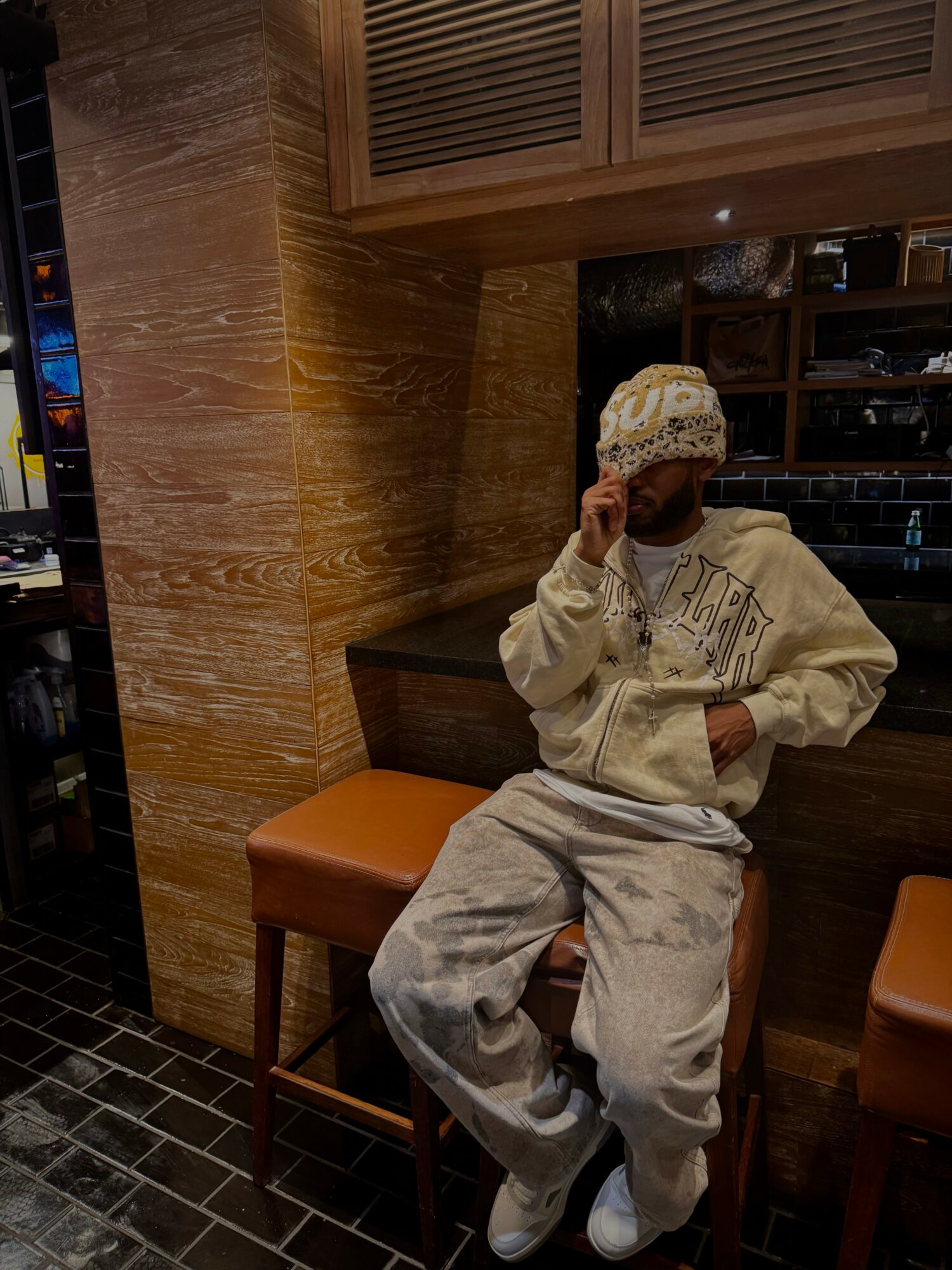 Person sitting on a barstool in a cafe, wearing a hoodie, sweatpants, and a beanie, with a hand near face.