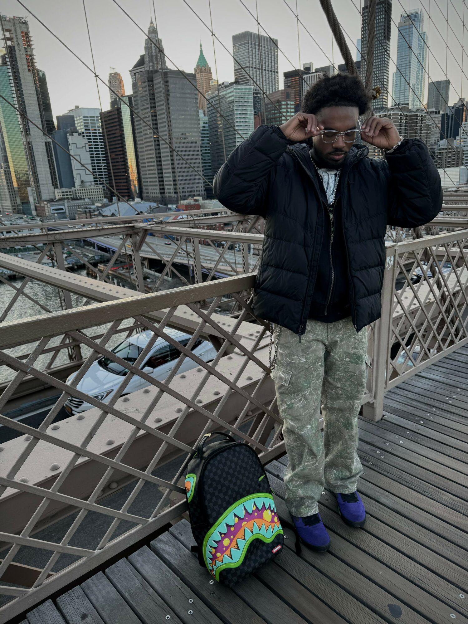 Man adjusting glasses on a bridge with city skyline in background, carrying a colorful backpack.