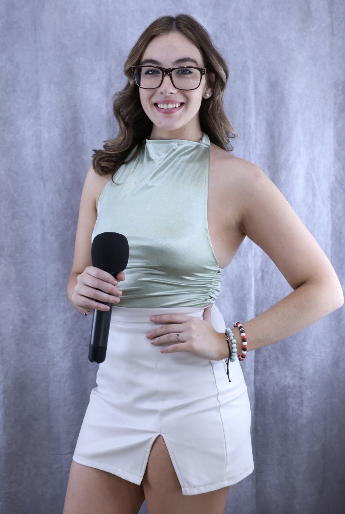 Young woman with glasses holding a microphone, smiling, wearing a sleeveless top and white skirt, standing against a gray background.