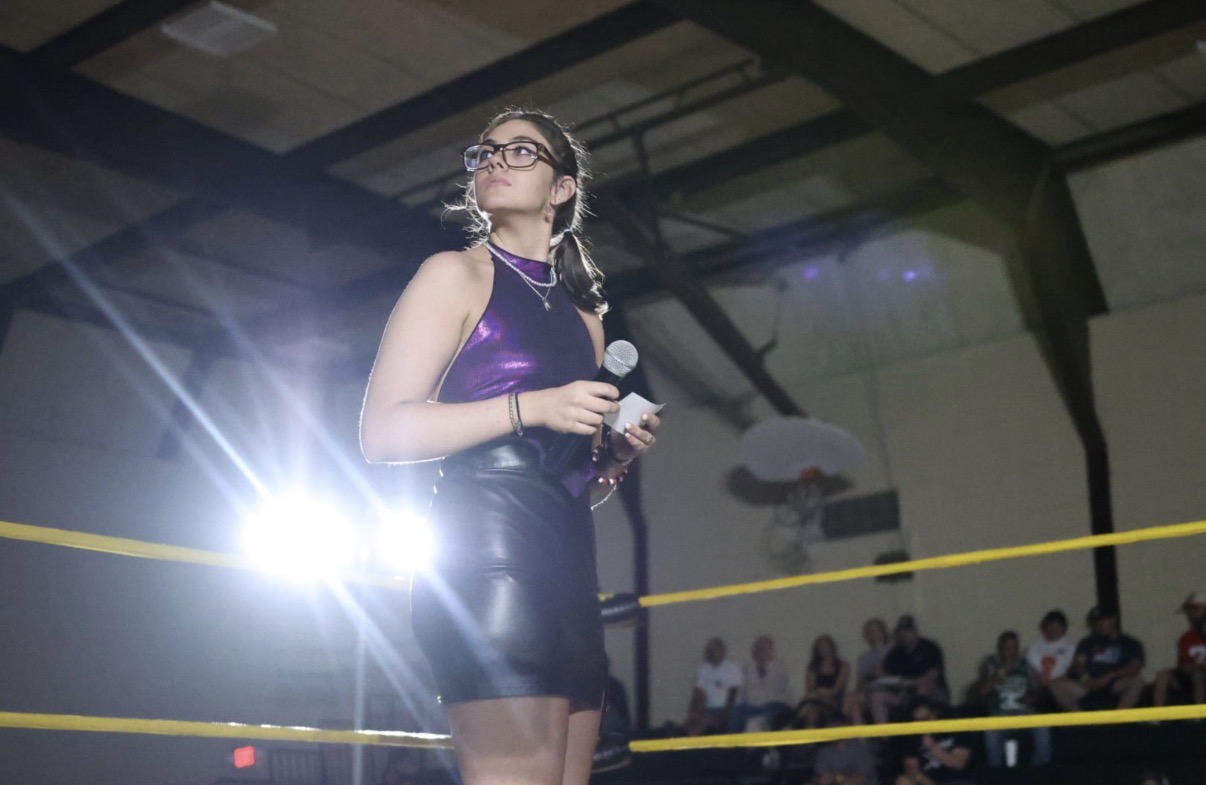 Woman in a purple dress holding a microphone in a sports arena with audience in background.