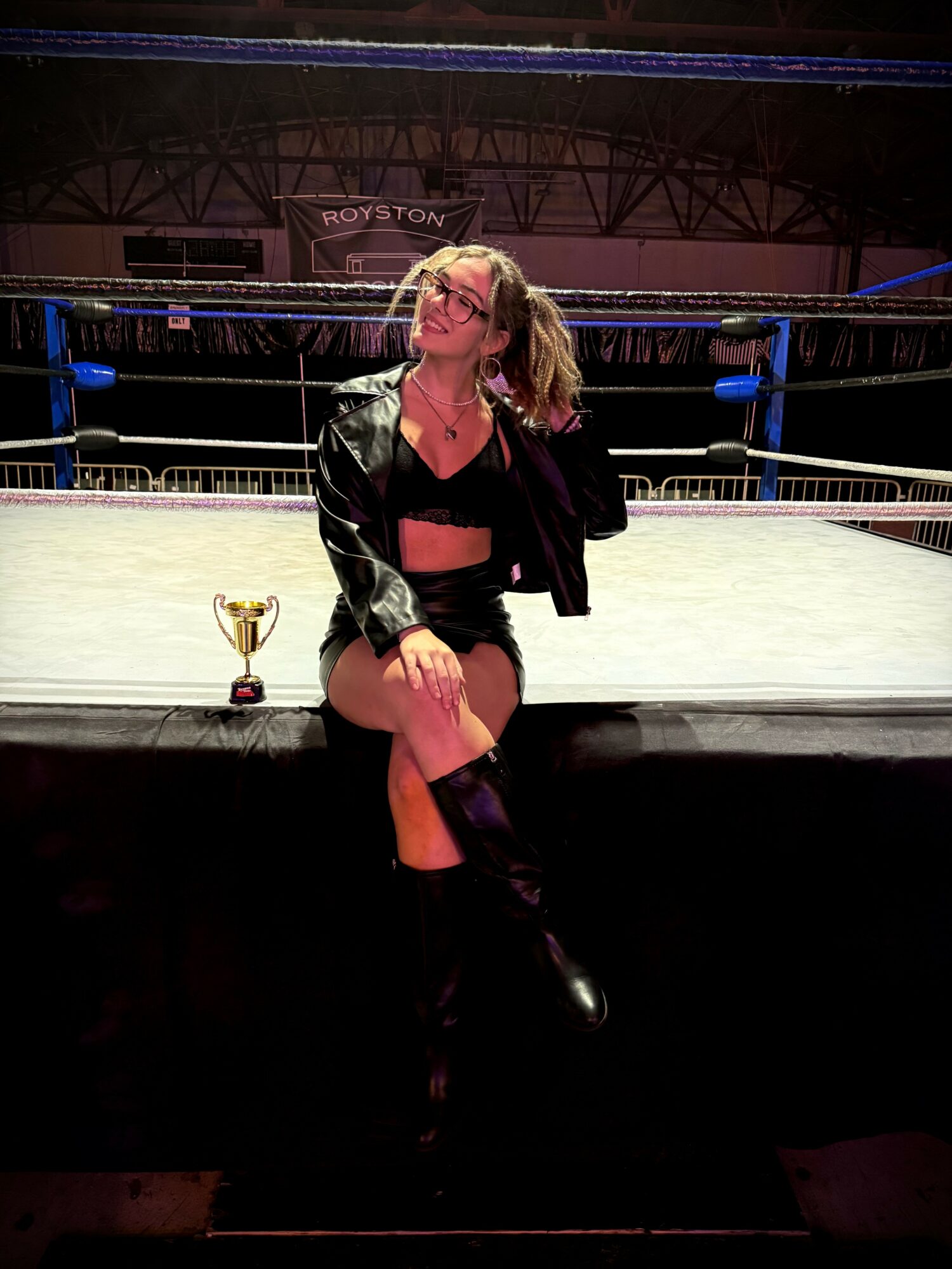 Person sitting on the edge of a wrestling ring with a trophy, in a large indoor arena.