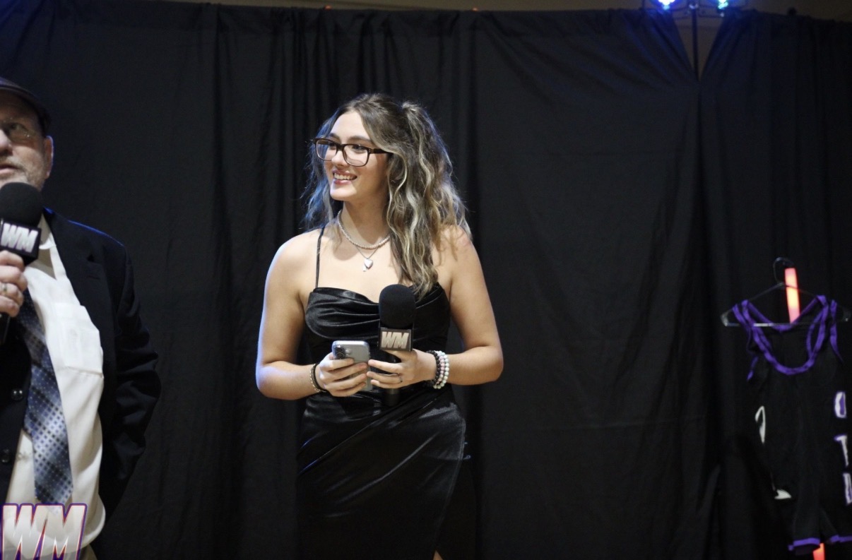 Woman with glasses in a black dress holding a microphone, standing on stage with a dark curtain background.