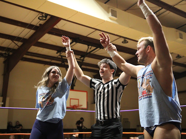 Two female volleyball players and a referee celebrate with raised hands in an indoor gymnasium.