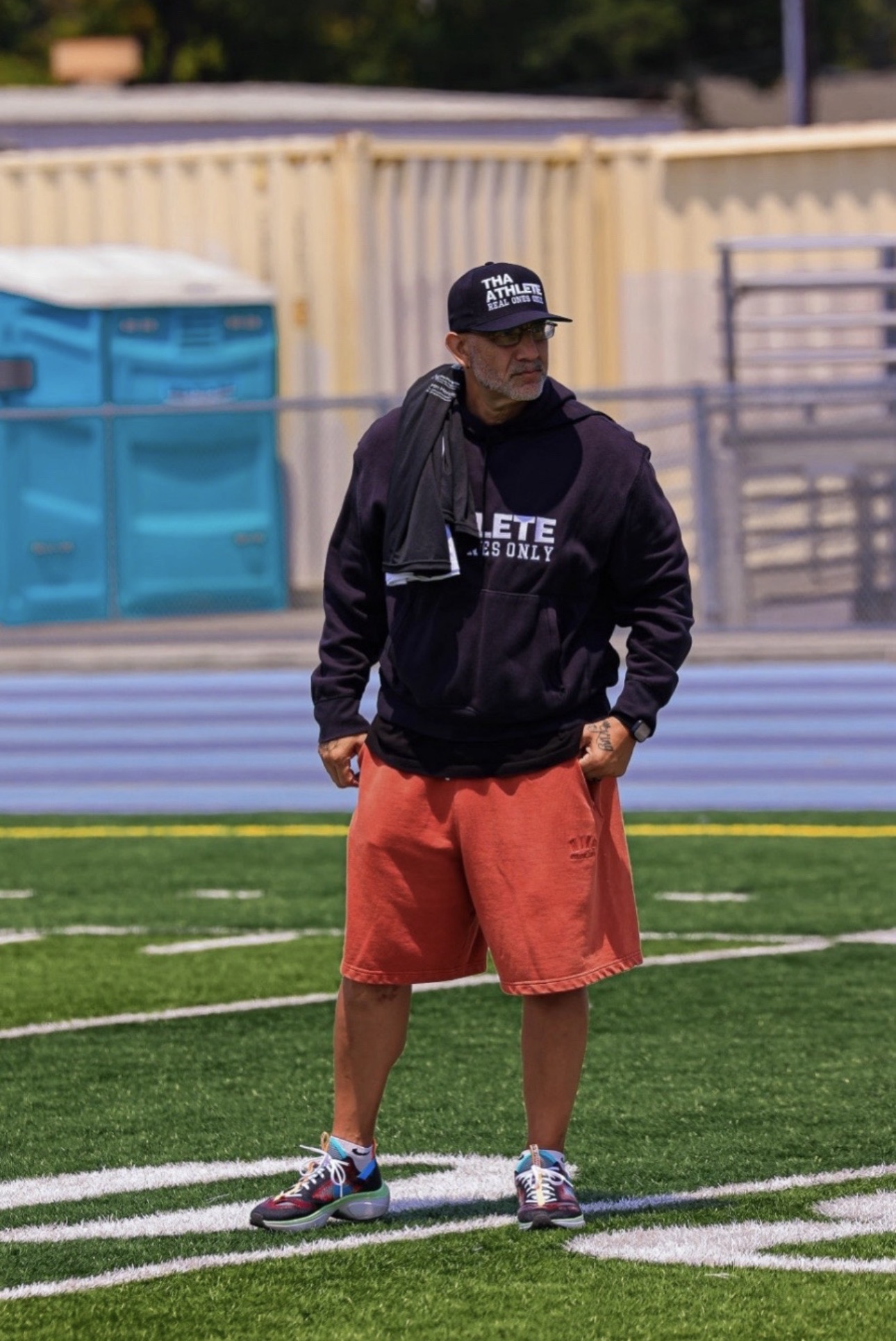 Man standing on a sports field wearing a black hoodie, red shorts, and a black cap, with a fence and blue recycling bins behind him.