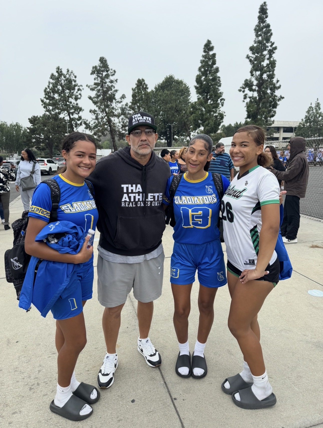 Four people standing outdoors, three young women in sports uniforms and one man in casual clothing, smiling.