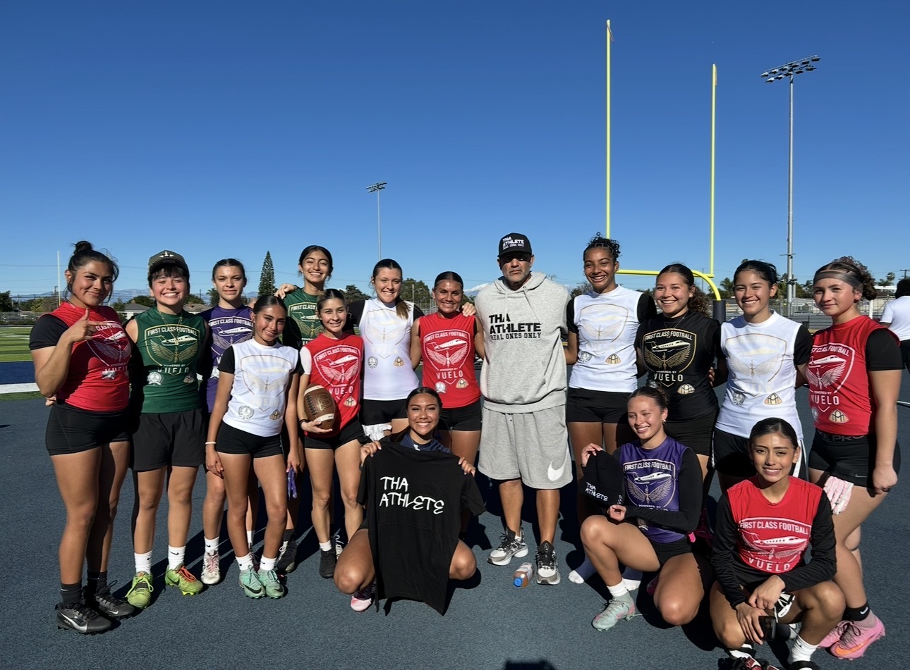 Group of young girls and a coach on a sports field with goalposts, smiling and posing for a photo.