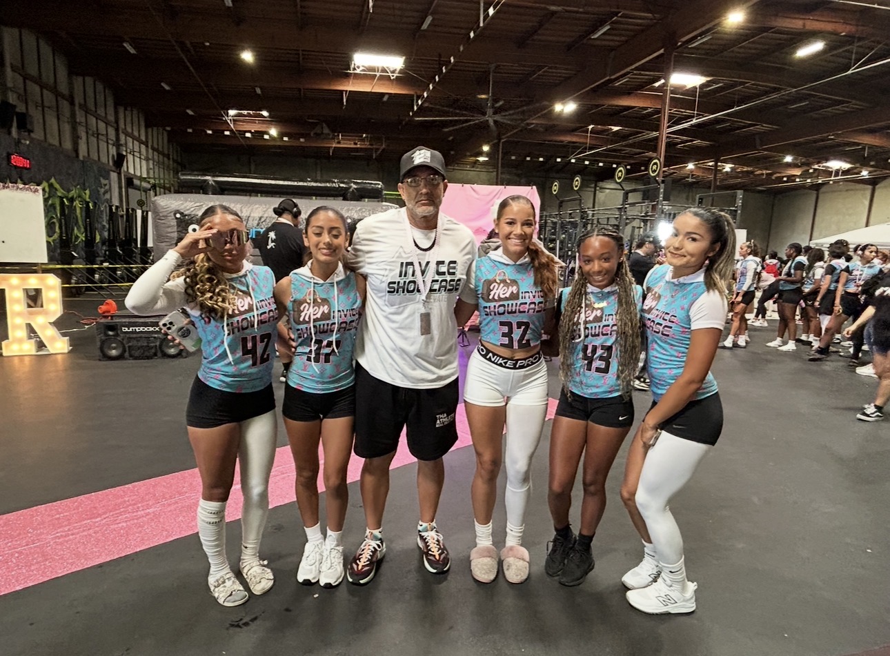 Group of six young female athletes and a male coach standing together indoors, smiling for a photo.