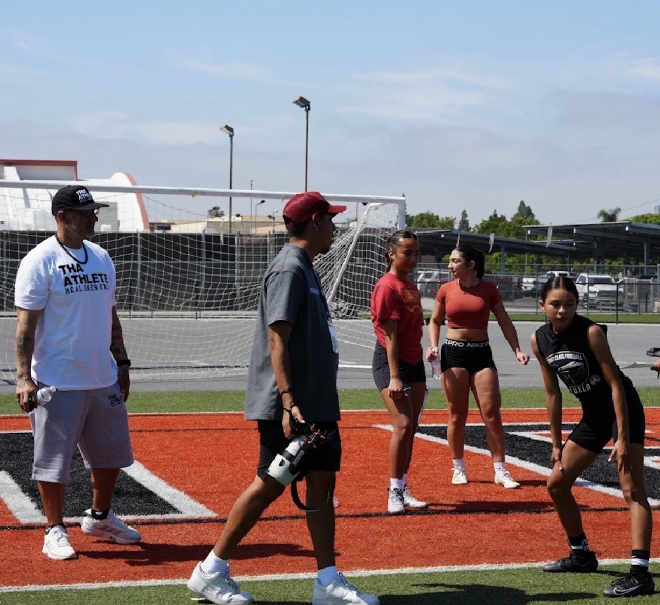Five people standing on a running track near a soccer goal, outdoors on a sunny day.