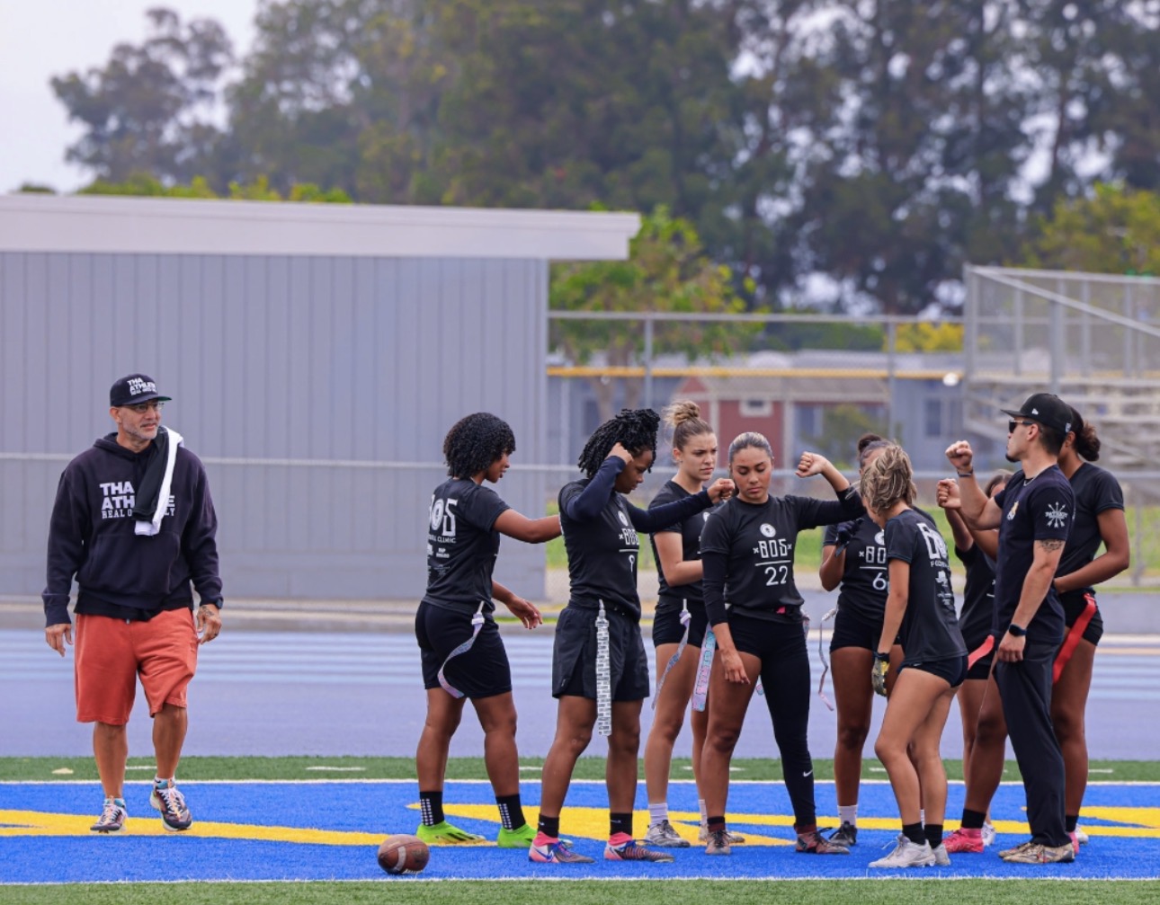 Group of women on a sports field, some celebrating, others talking, with a man in shorts and a hoodie nearby.