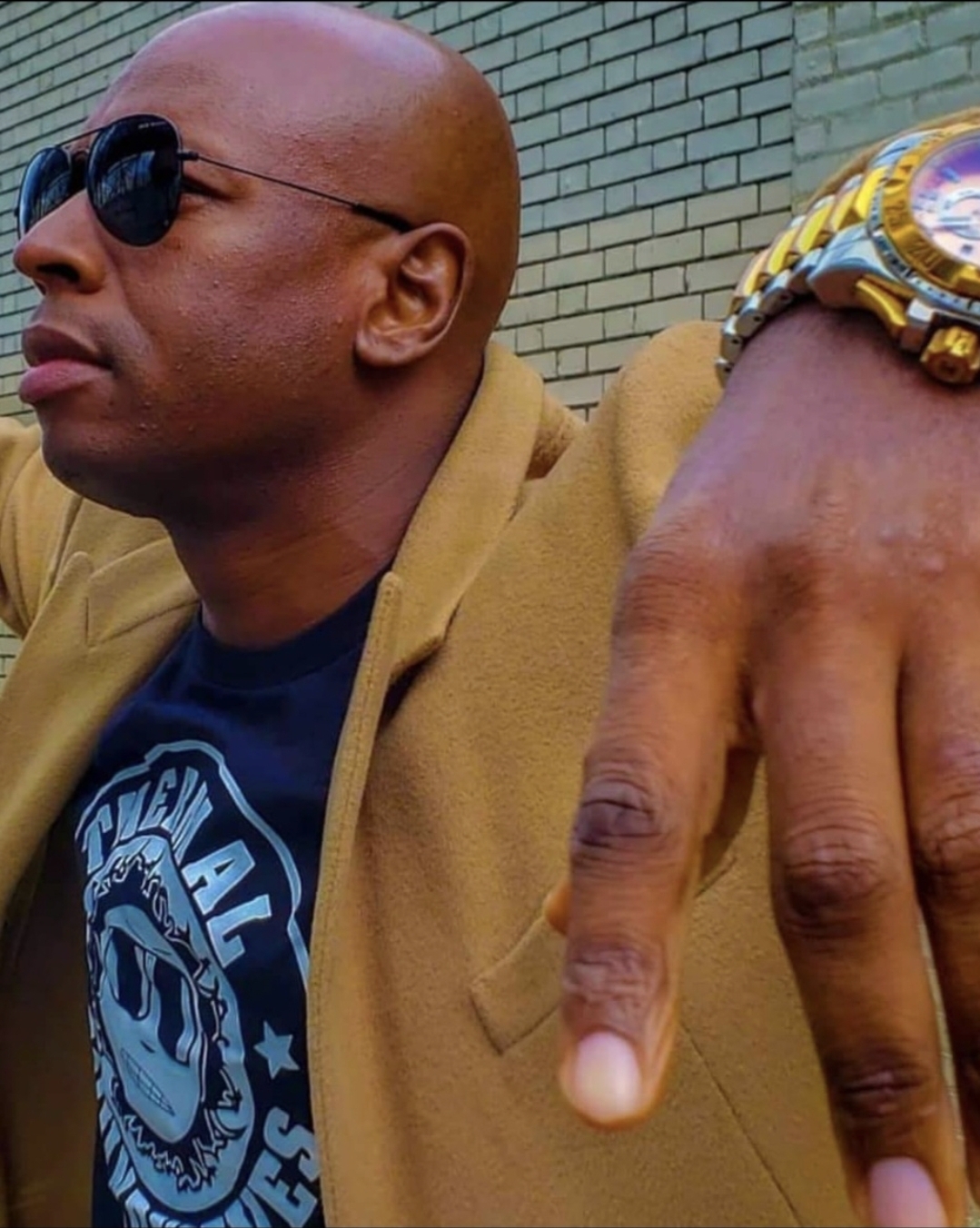 Bald man wearing sunglasses and a tan jacket, with a hand showing a watch and pointing finger.