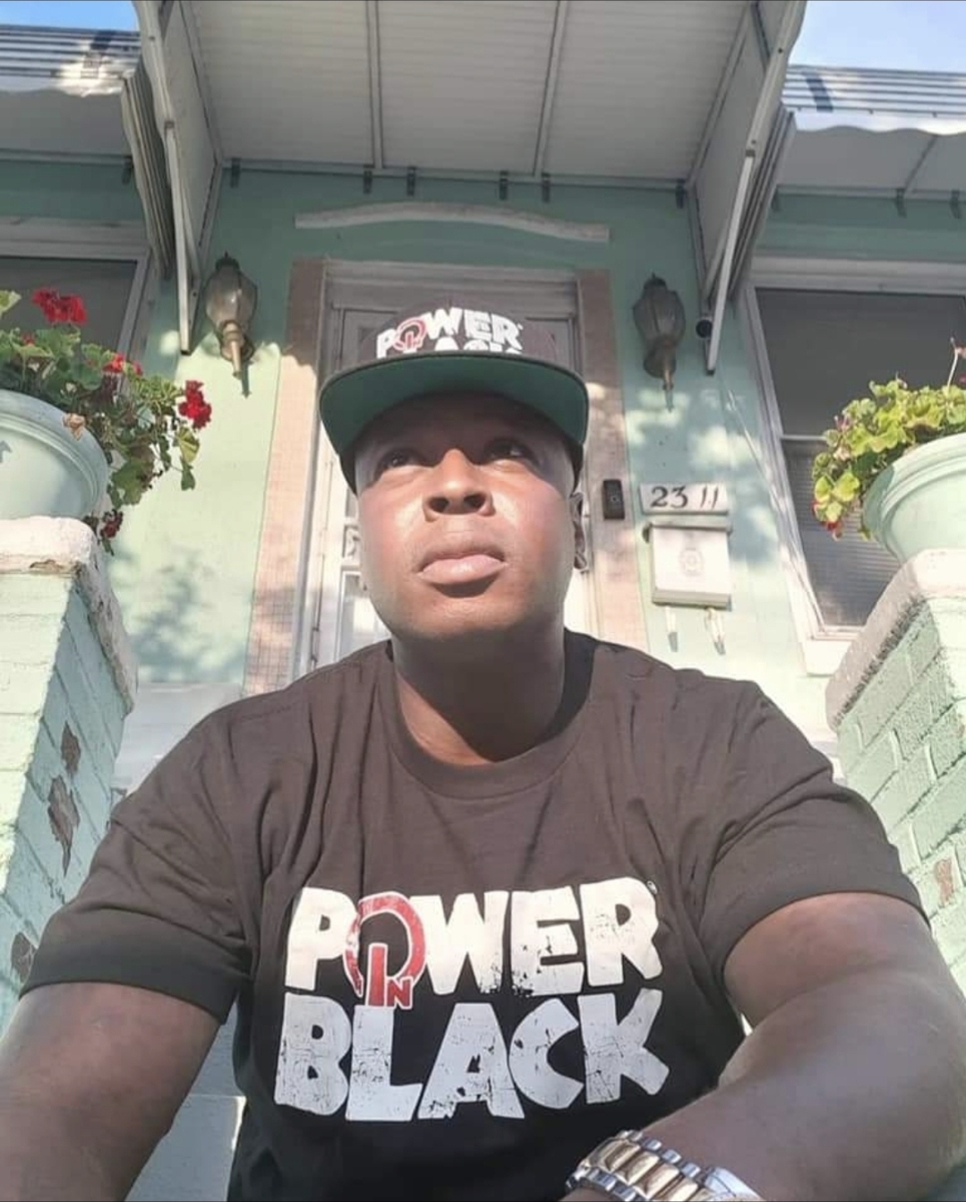 Young man wearing a black T-shirt and cap, sitting outside in front of a house with potted plants, sunlight on face.