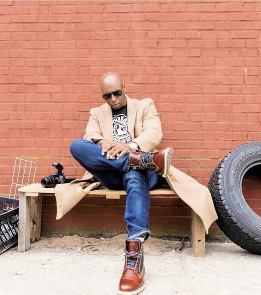 Man sitting on a wooden bench against a red brick wall, wearing sunglasses, beige coat, jeans, and boots.