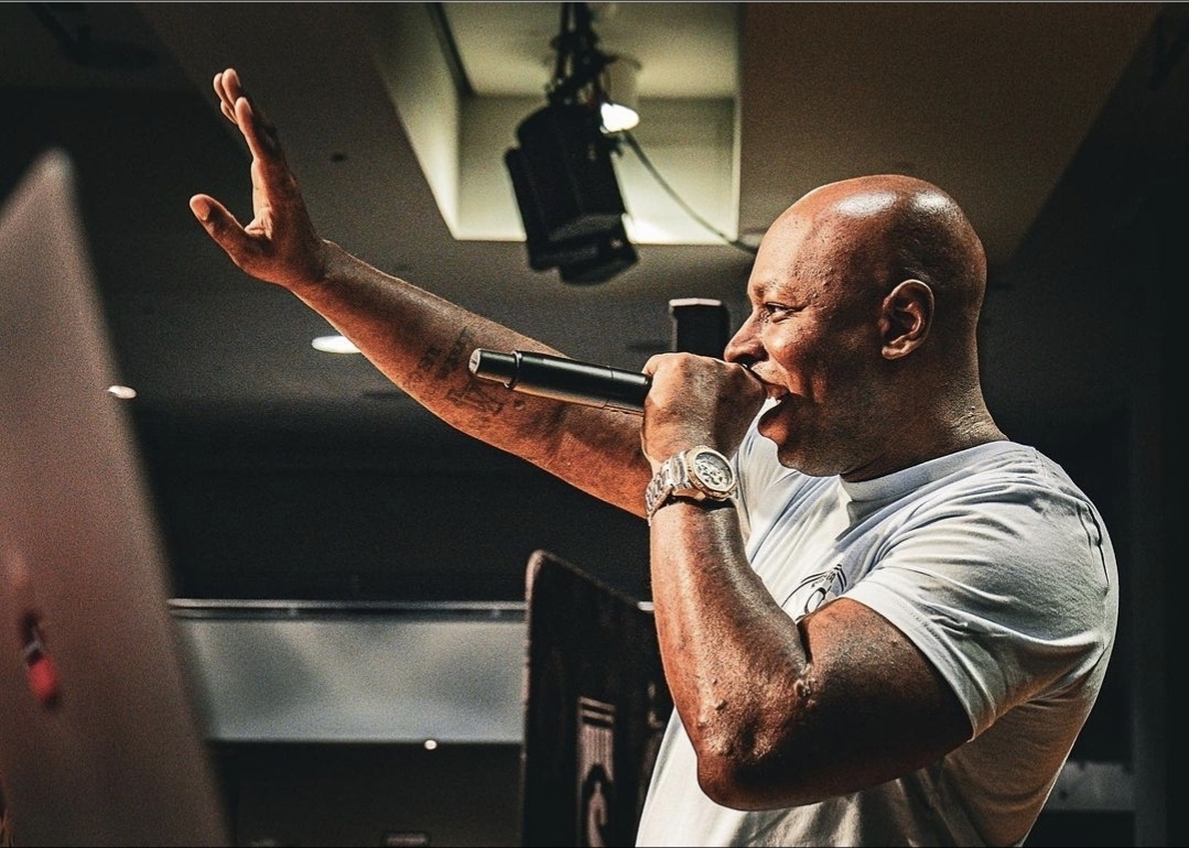 Man holding microphone, gesturing with his right hand, in a dimly lit room with ceiling lights and equipment.