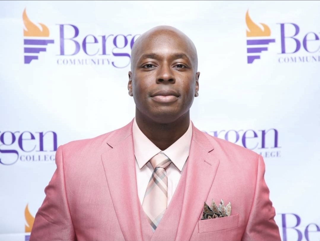 Man in pink suit and tie standing in front of Bergan College backdrop.