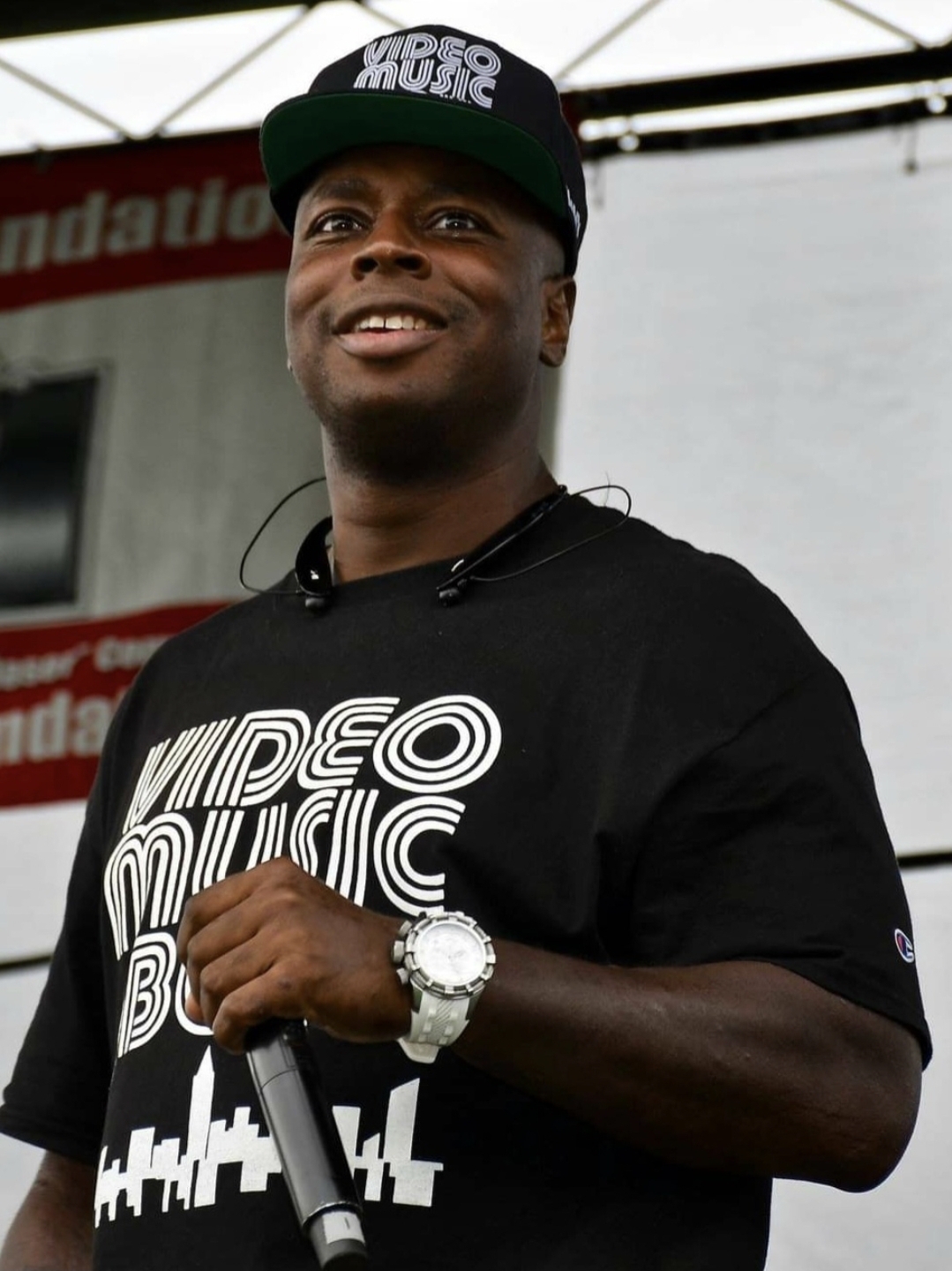 Man wearing a black cap and T-shirt holding a microphone, smiling outdoors.