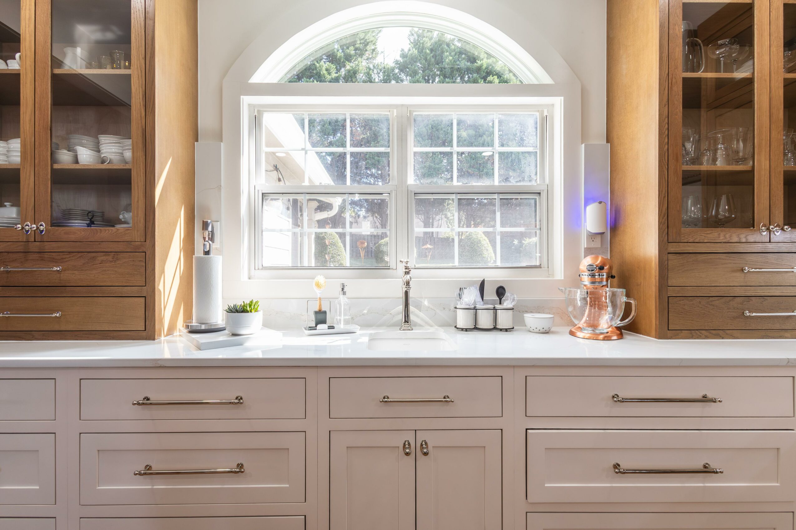 Kitchen window with white frame and arched top, view of trees outside, surrounded by wooden cabinets and a white countertop.