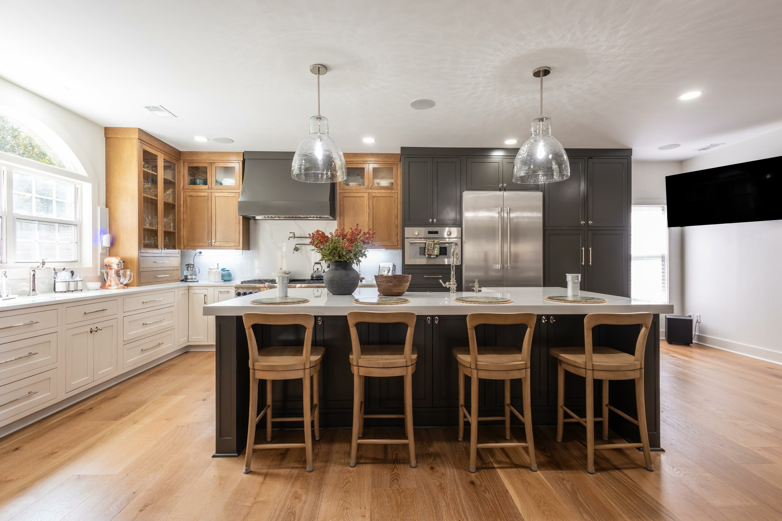Modern kitchen with black and wooden cabinets, island with five chairs, stainless steel appliances, and pendant lights.