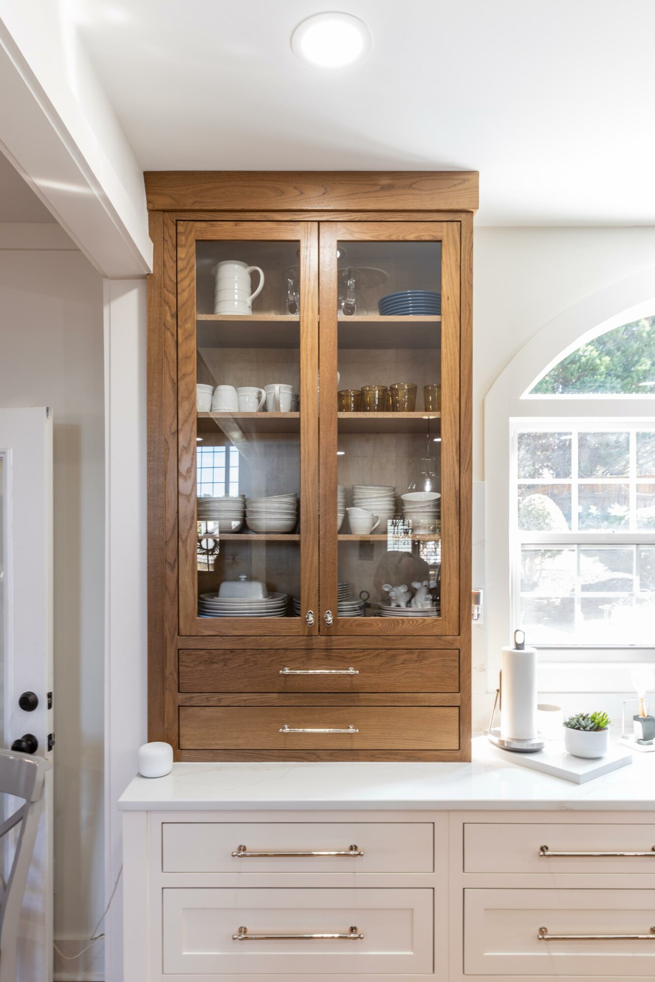 Tall wooden cabinet with glass doors displaying dishes and glassware, positioned next to a window in a bright room.