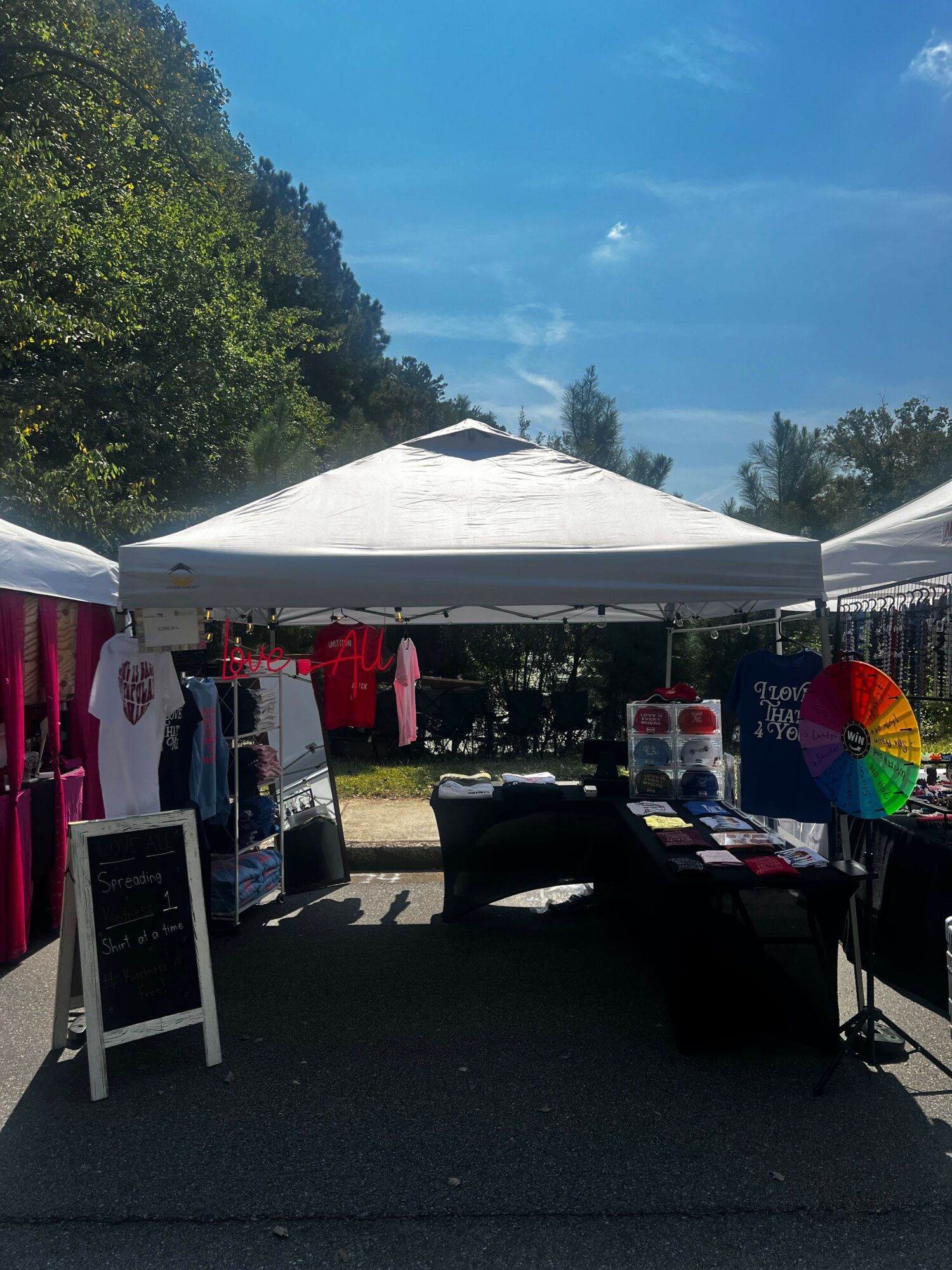 Outdoor market stall with tents, clothing, and a rainbow umbrella, set against a blue sky and trees.