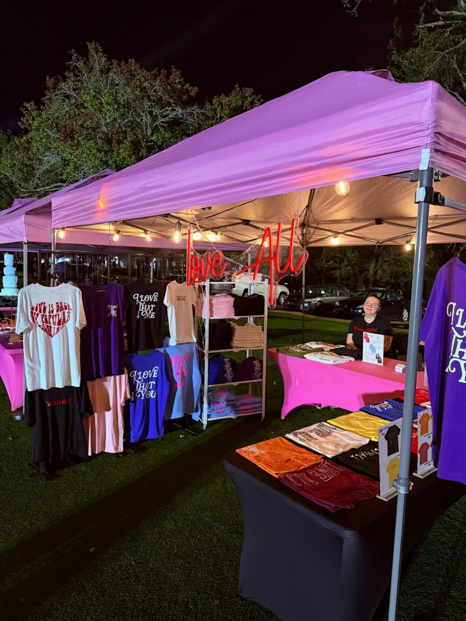 Outdoor market stall with clothing and accessories under a pink canopy at night.