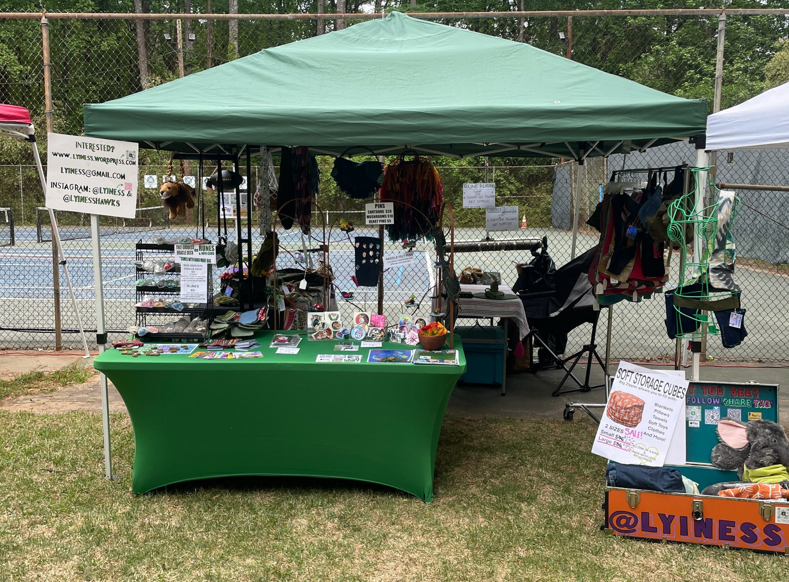 Outdoor market stall with a green canopy, table displaying items, and a fenced background with trees.