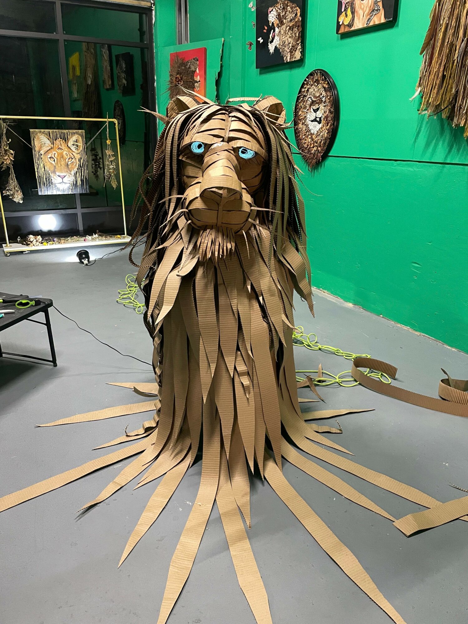 Large lion sculpture made of layered paper or cardboard with blue eyes, positioned indoors against a green wall with artwork.